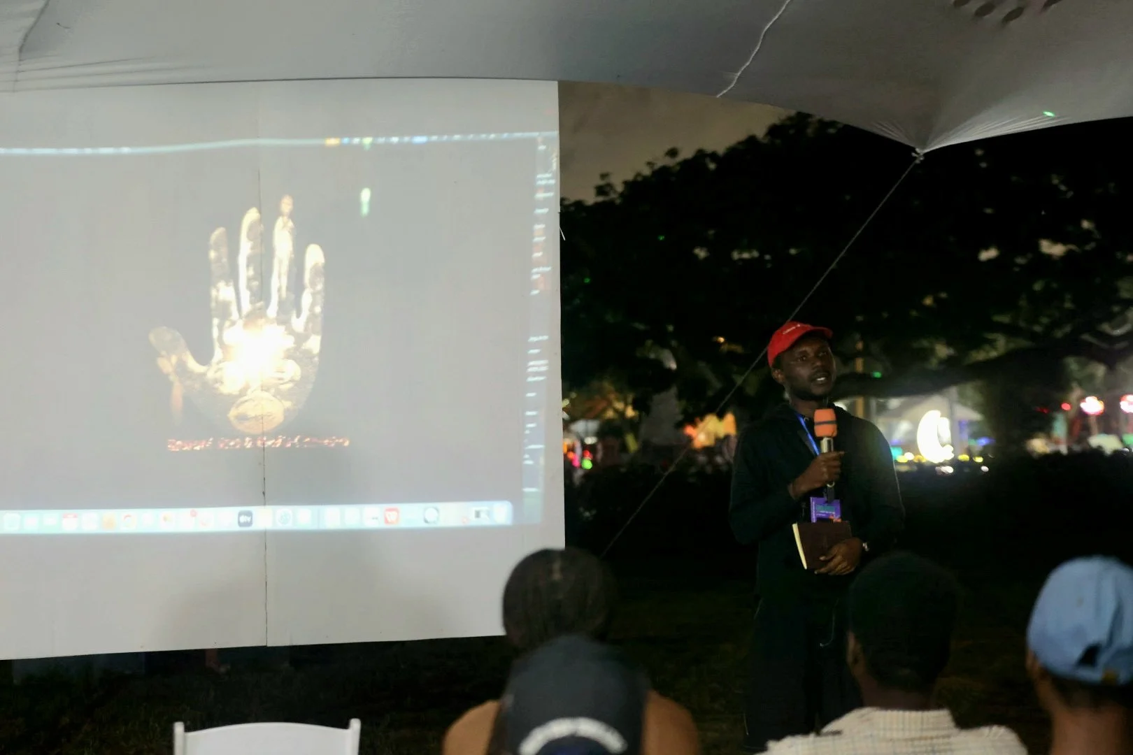 Person speaking into microphone during outdoor event at night, with a projection of a robotic hand on a large white screen behind them, and audience members seated in front.