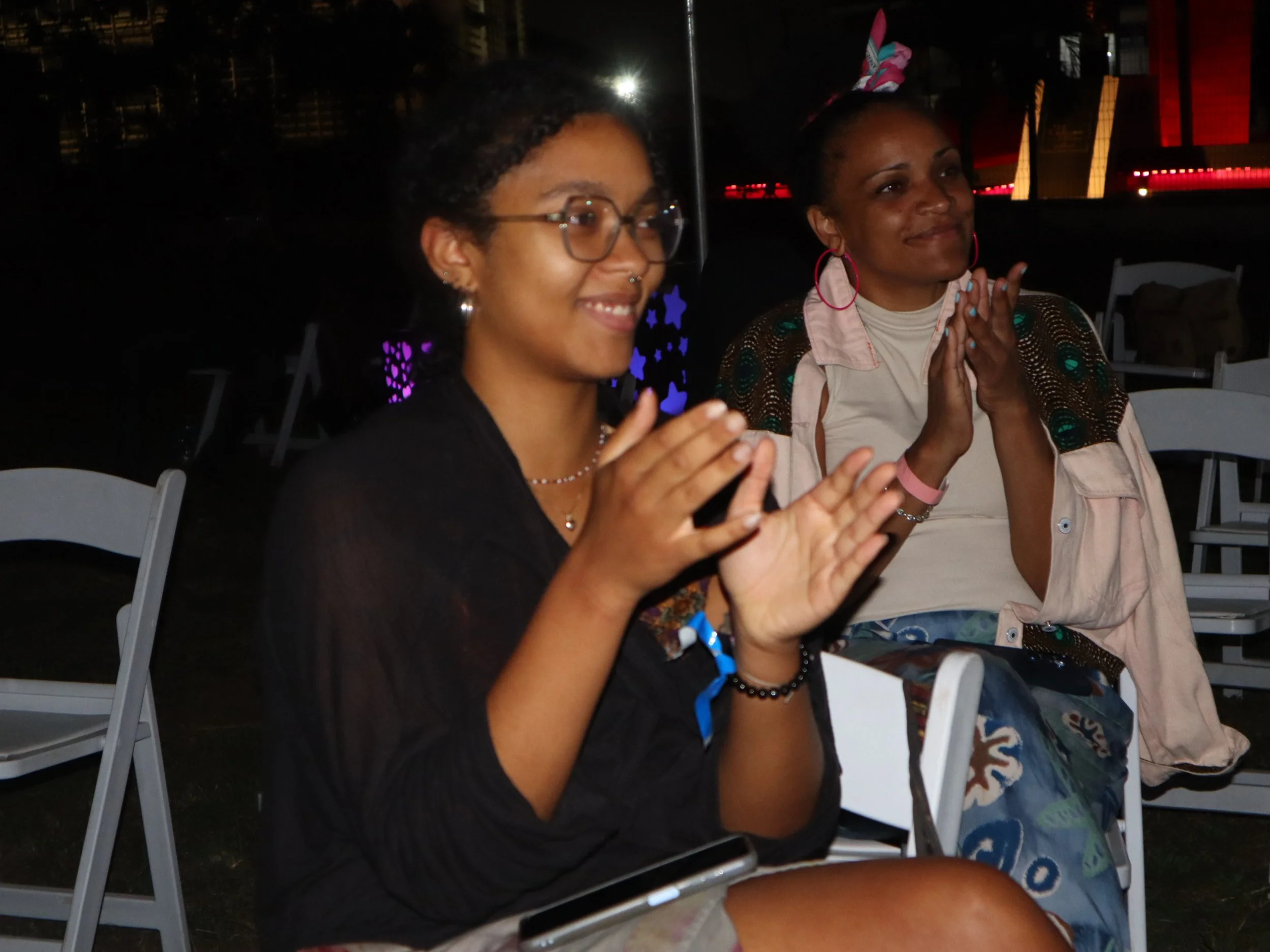 Two women sitting at an outdoor event during nighttime, smiling and clapping, one wearing glasses and earrings, the other with hoop earrings and a patterned shirt.