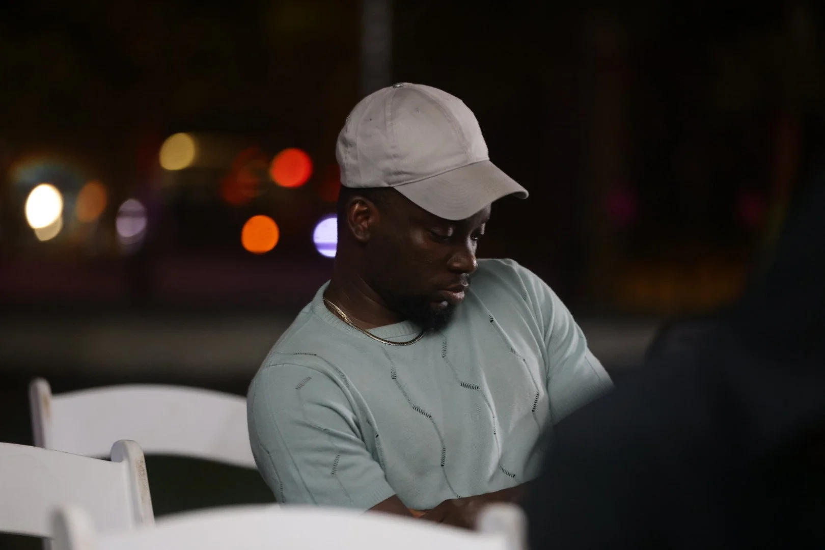 A young man with a cap and gold chain sitting outdoors at night, looking down