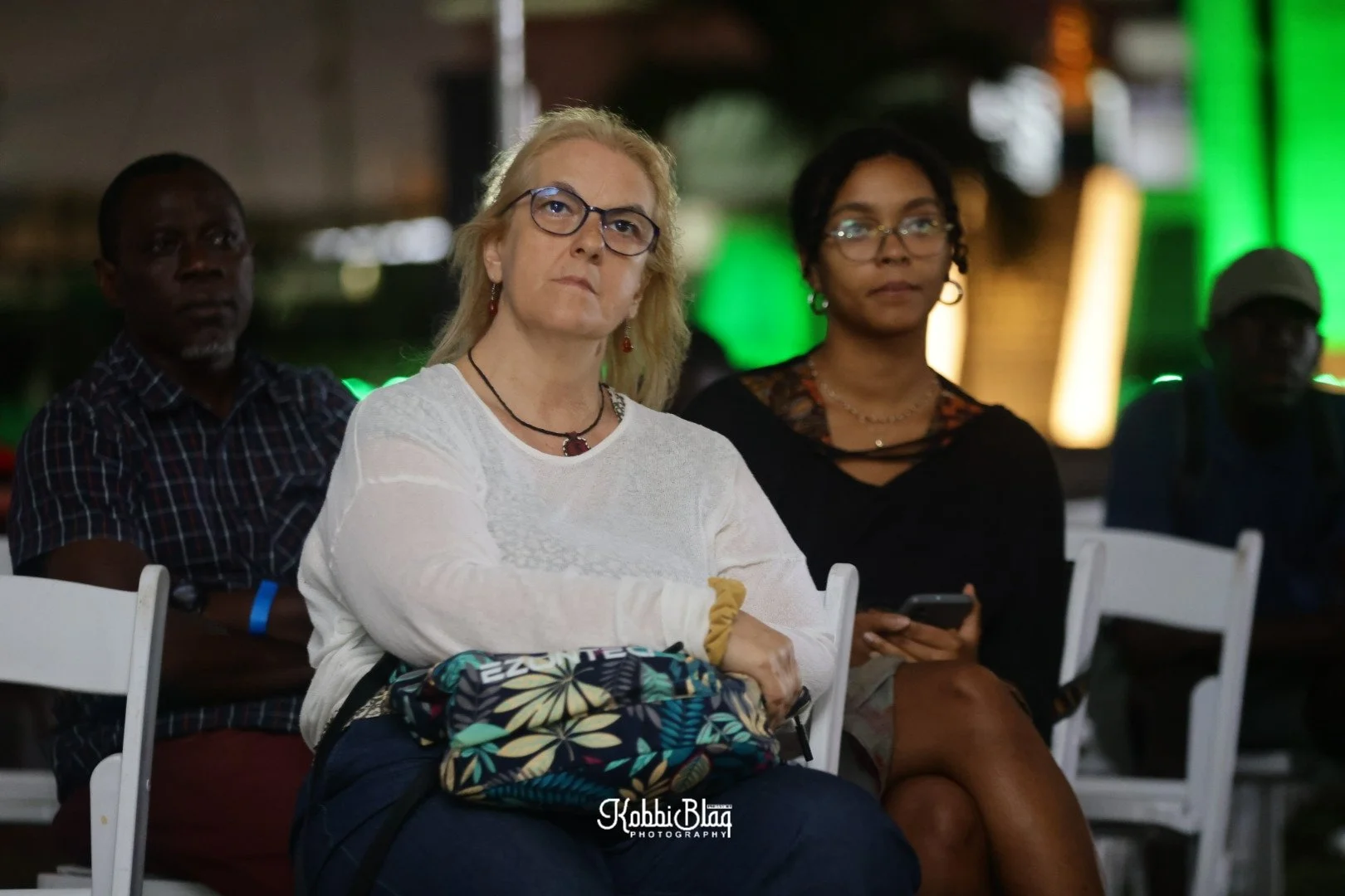 A group of people sitting on white chairs at an indoor event, with two women in the foreground, one with blonde hair and glasses wearing a white top, and the other with black hair and glasses wearing a black top, both looking attentive. There are men