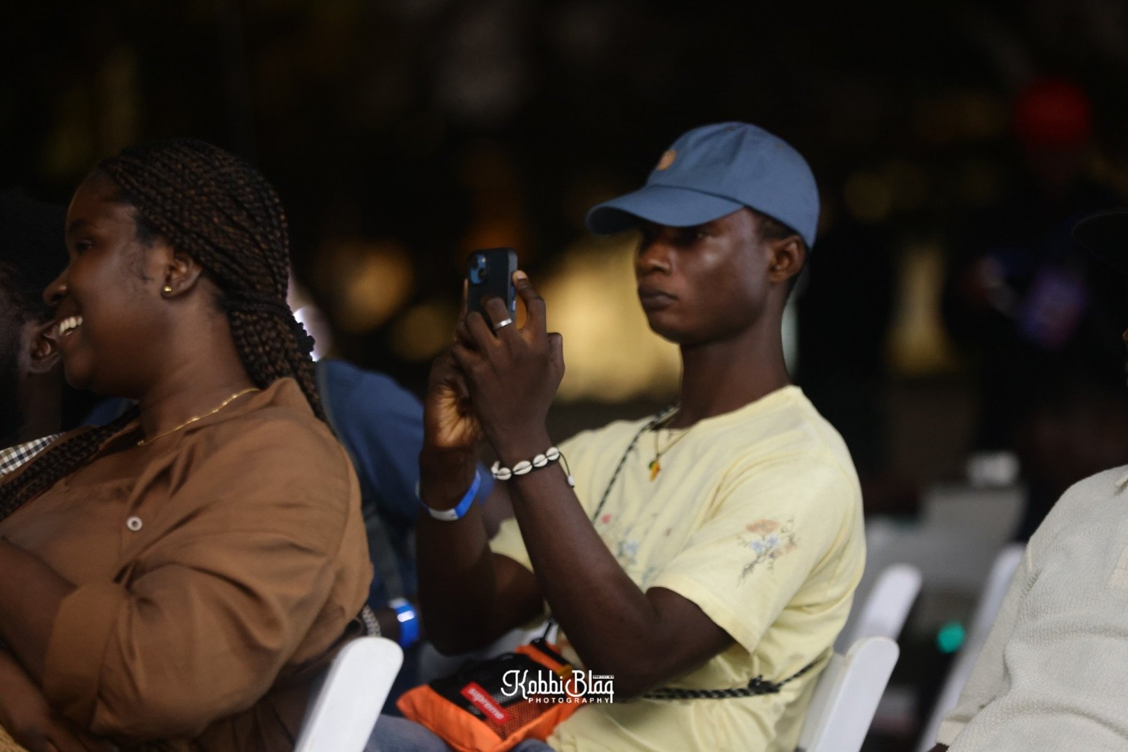 A young man in a blue cap and light yellow shirt is sitting and holding a phone, appearing to take a photo or video. Next to him is a smiling woman with braided hair, wearing a brown shirt. They are seated in chairs at an indoor event with a dark bac