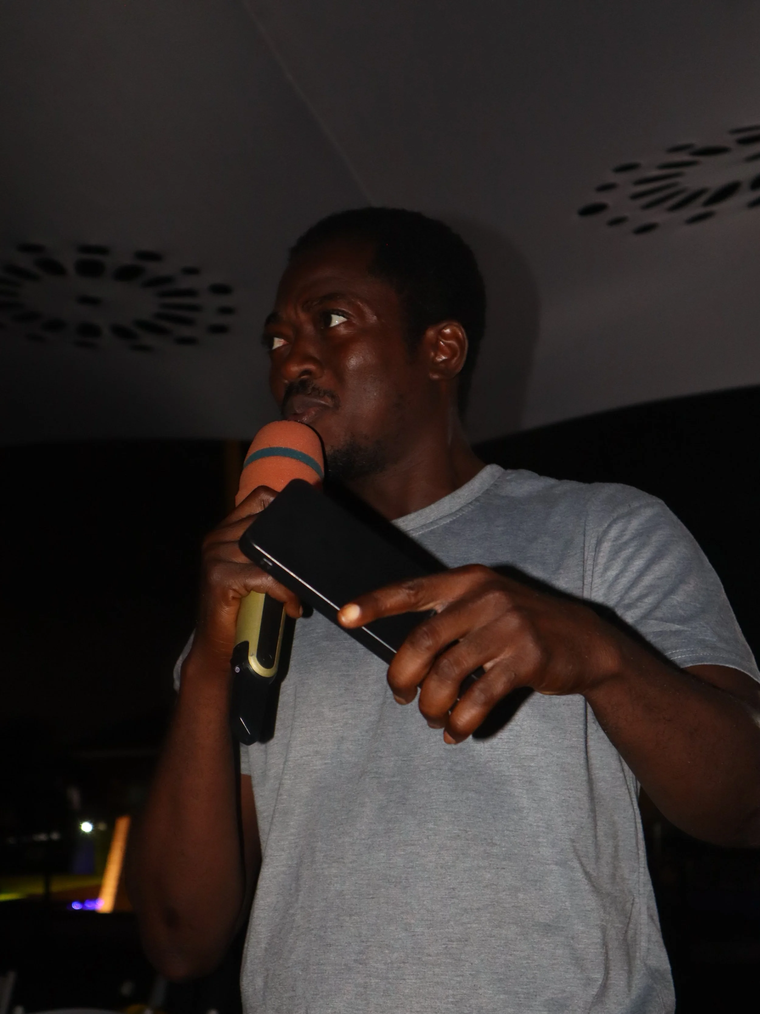 A man holding a microphone and a smartphone, wearing a gray t-shirt, in a dark environment.