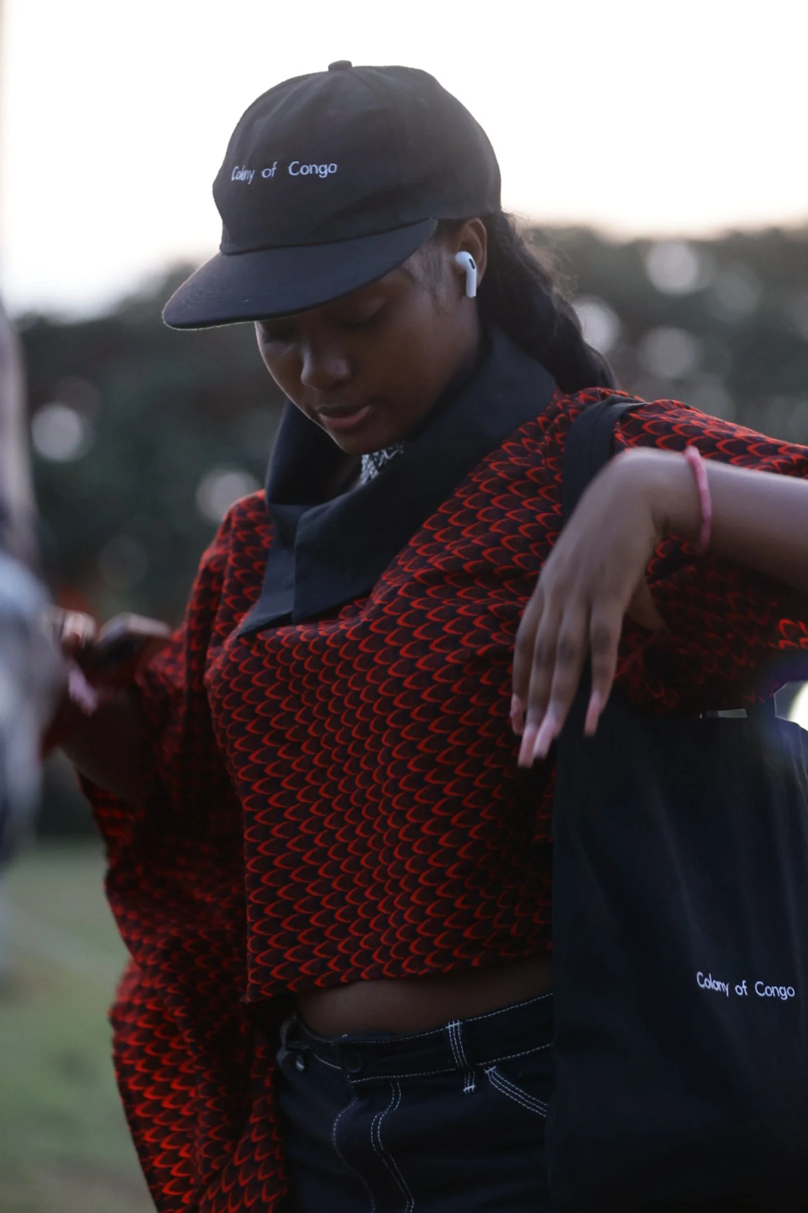A woman wearing a black cap with white text reading 'Colony of Congo,' white wireless earbuds, and a black and red patterned top, with her head tilted downward and her arm raised, outdoors with a blurred background of trees and sky.