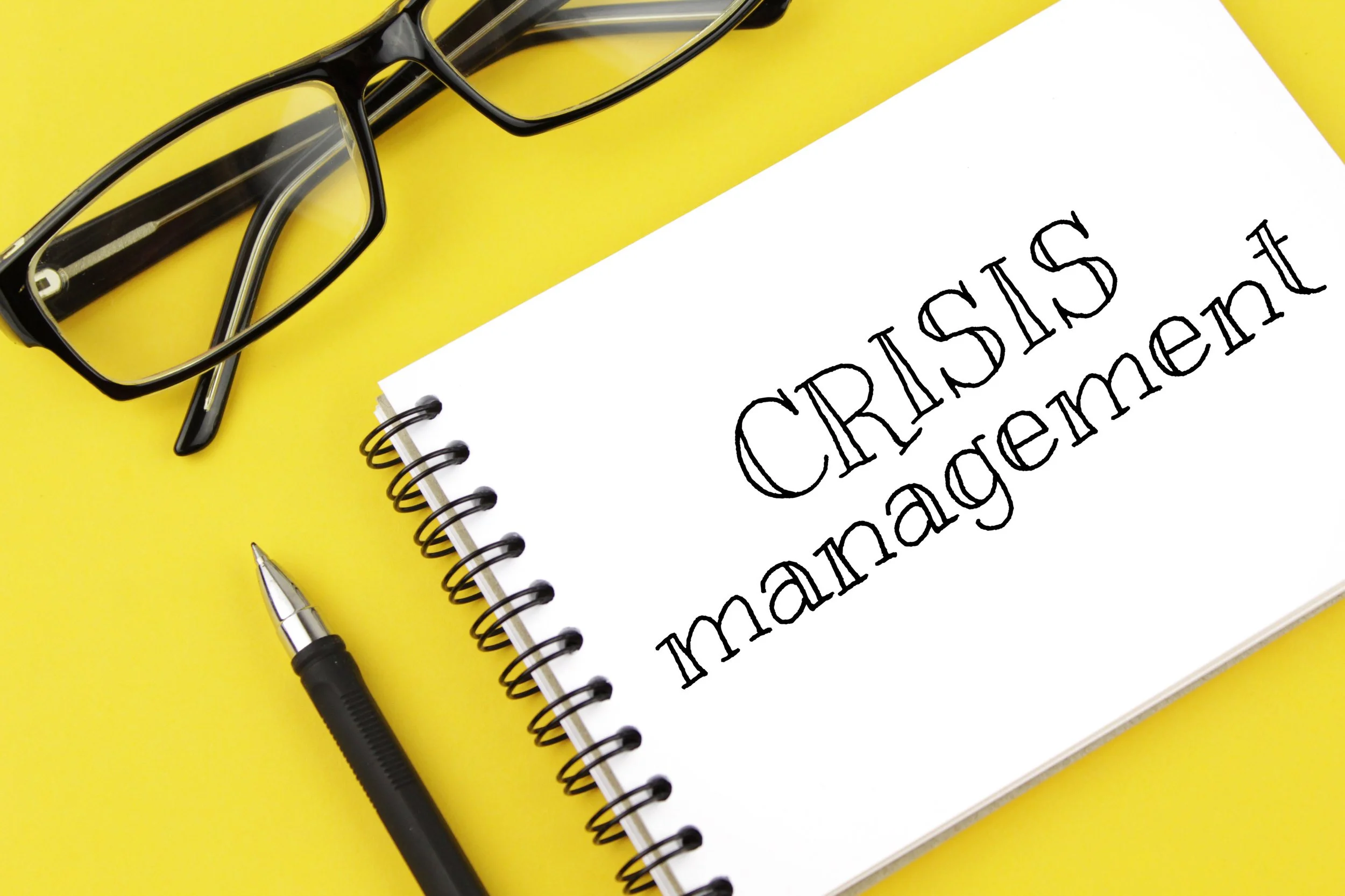 A pair of black glasses, a black pen, and a spiral notebook with the words 'CRISIS management' written on a white page, all on a yellow background.