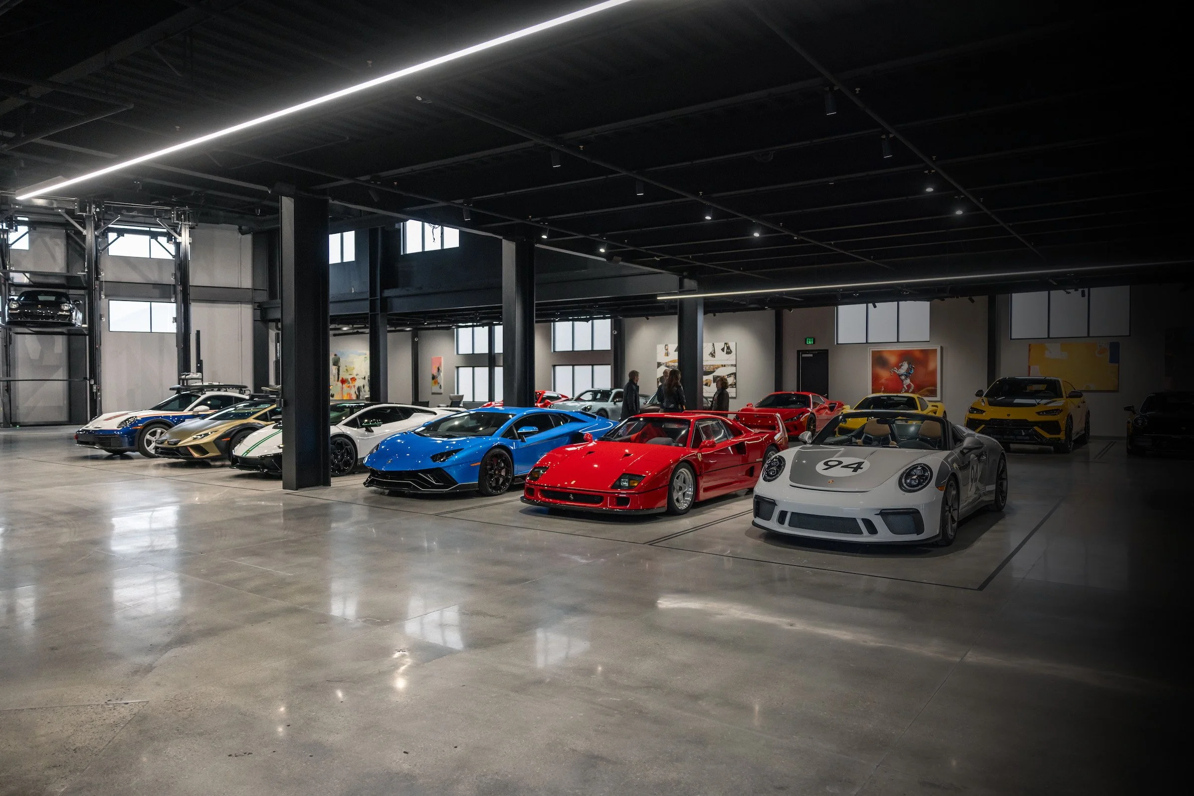 Luxury car showroom with a collection of high-end sports cars, including Lamborghini, Ferrari, Porsche, and others, displayed in a spacious, modern interior with polished concrete floors and large windows.