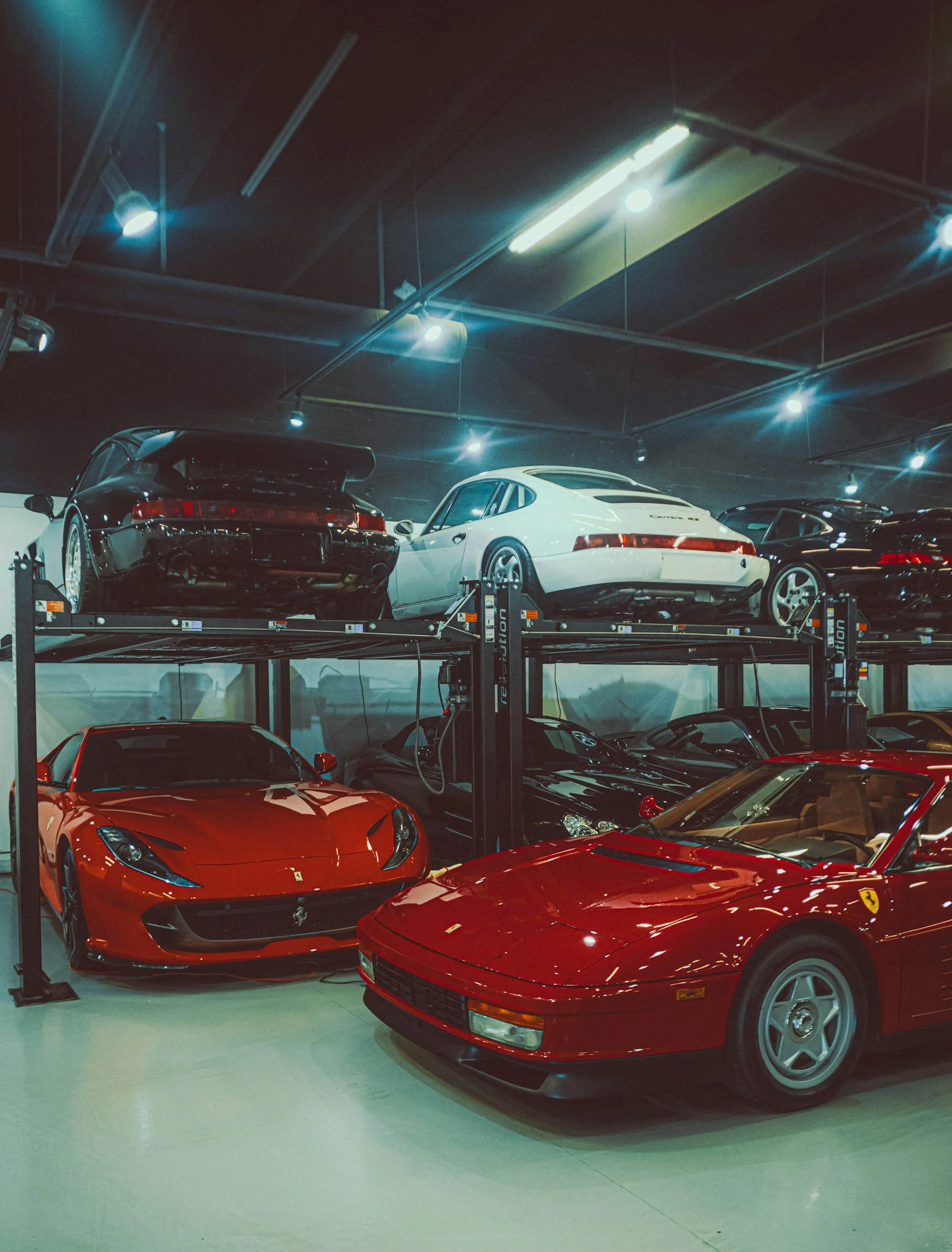 A collection of high-end sports cars, including a red Ferrari and a red Porsche, stored in a luxury indoor garage with a second level of parked cars.