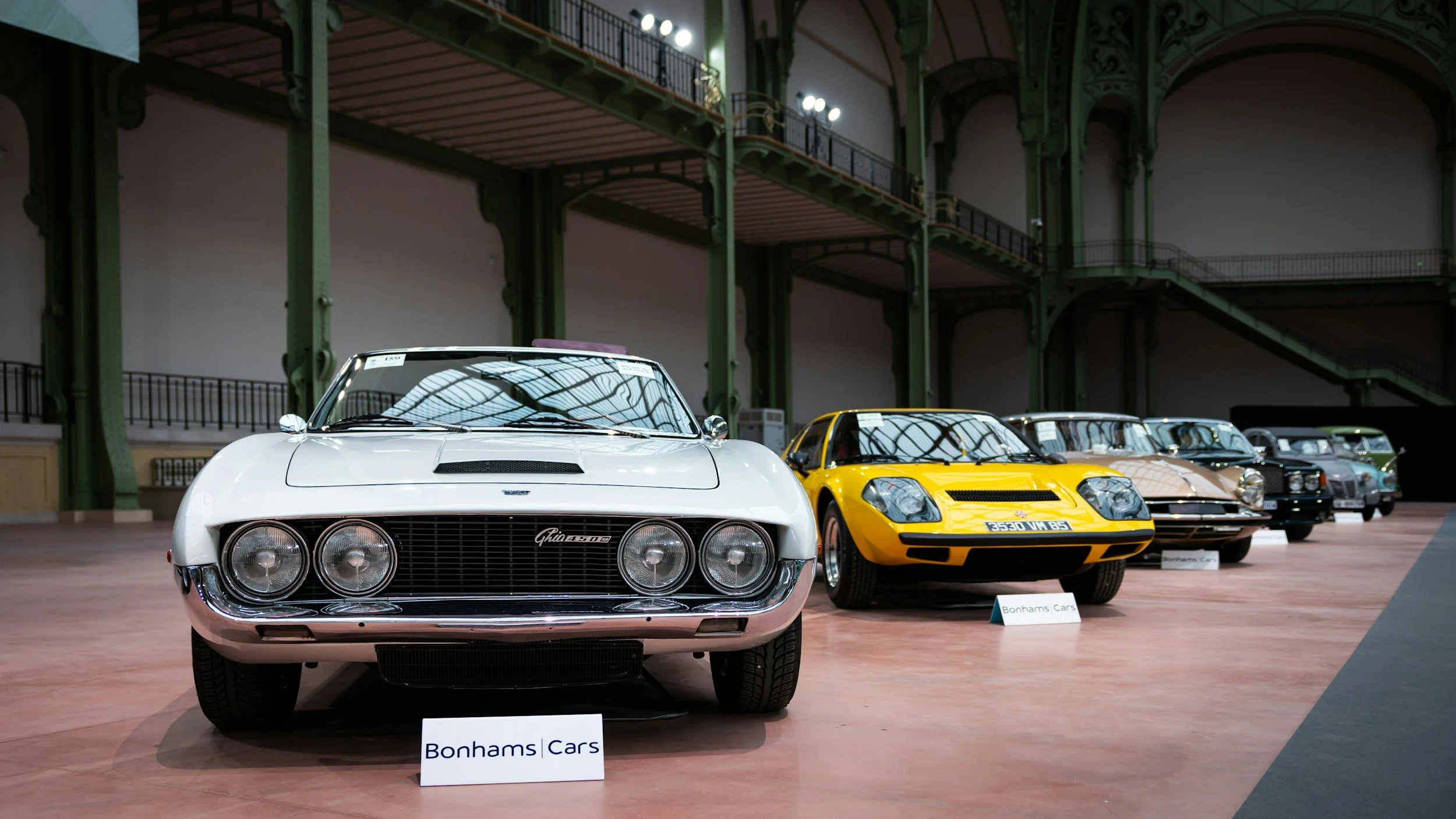A lineup of classic cars displayed in an exhibition hall with ornate green metal architecture and wooden flooring, including a white Aston Martin with a sign reading Bonhams Cars in the foreground, a yellow sports car, and several other vintage vehicles.