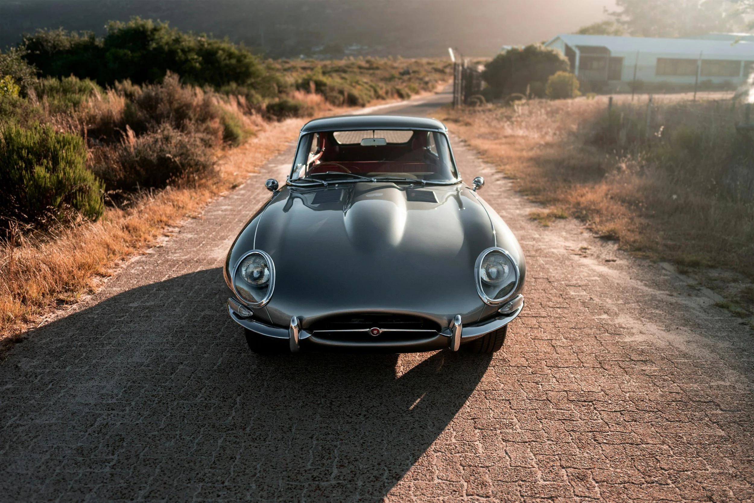 A vintage black sports car parked on a dirt road surrounded by dry grass and bushes, with a building and a fence in the background, during sunset.