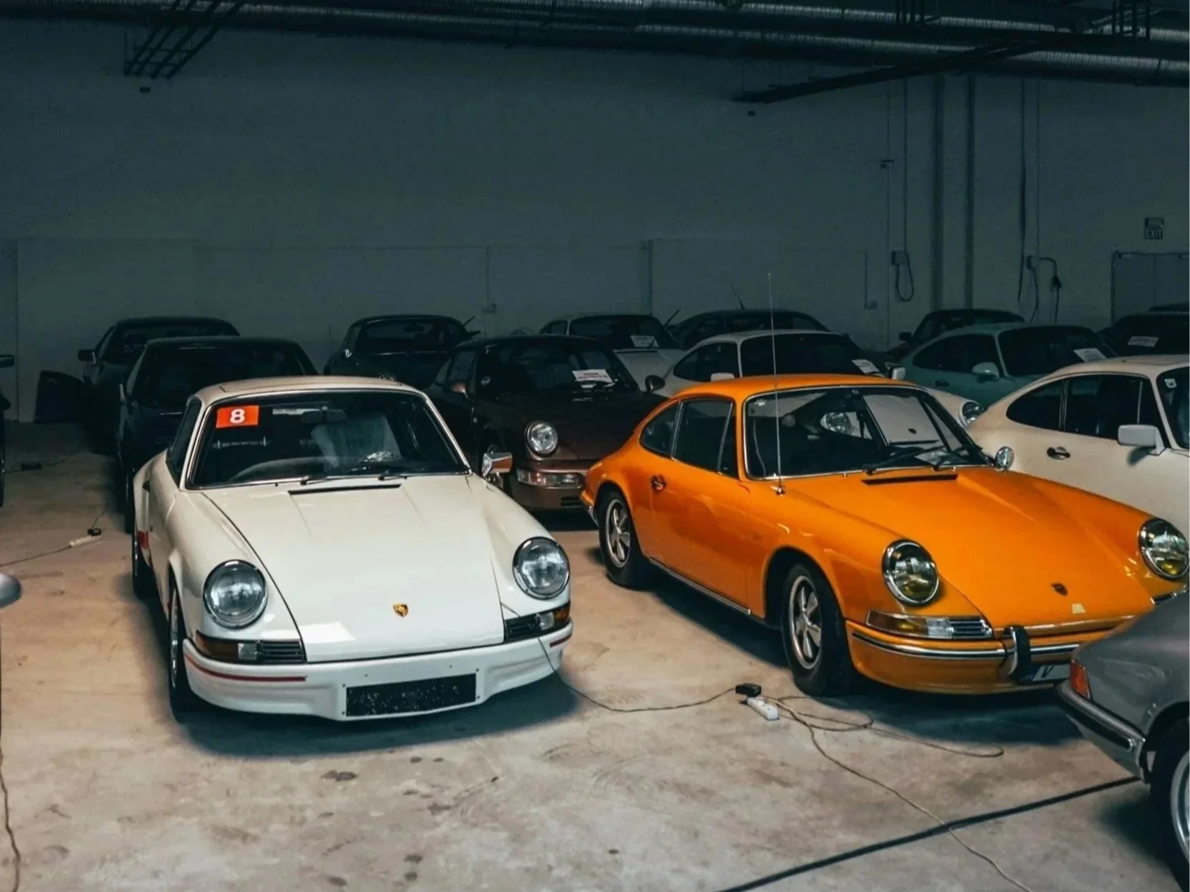 Several vintage Porsche cars inside a warehouse, including a white and an orange model in the foreground, surrounded by other cars.