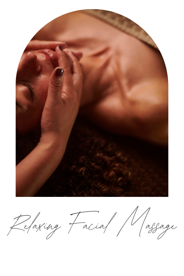 A person lying down with eyes closed, receiving a facial massage, with a relaxed expression and curly hair visible, in a warm-toned setting.