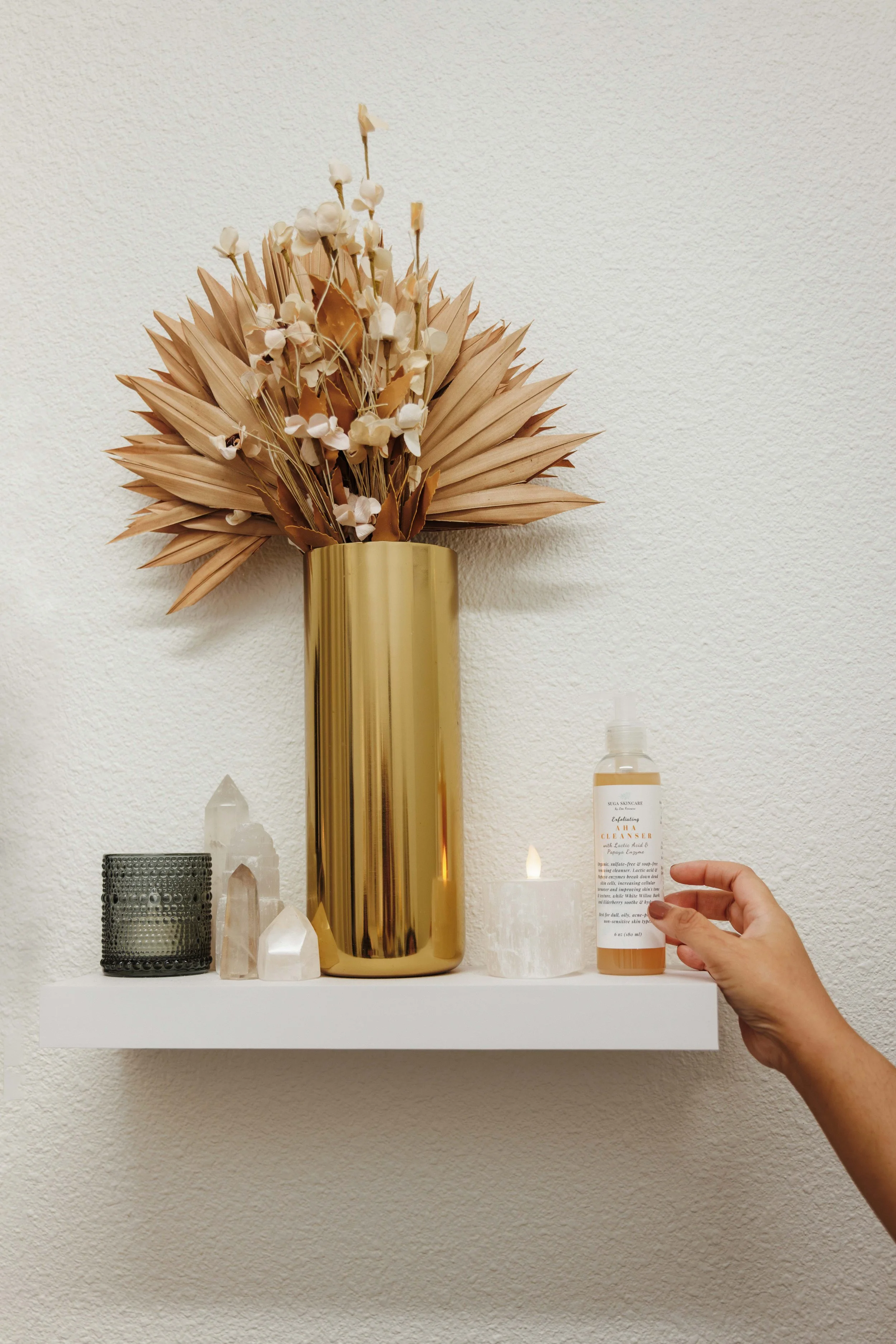 Decorative shelf with a gold vase holding dried flowers, a candle, a black textured container, a cluster of crystals, and a hand holding a spray bottle of facial cleanser.