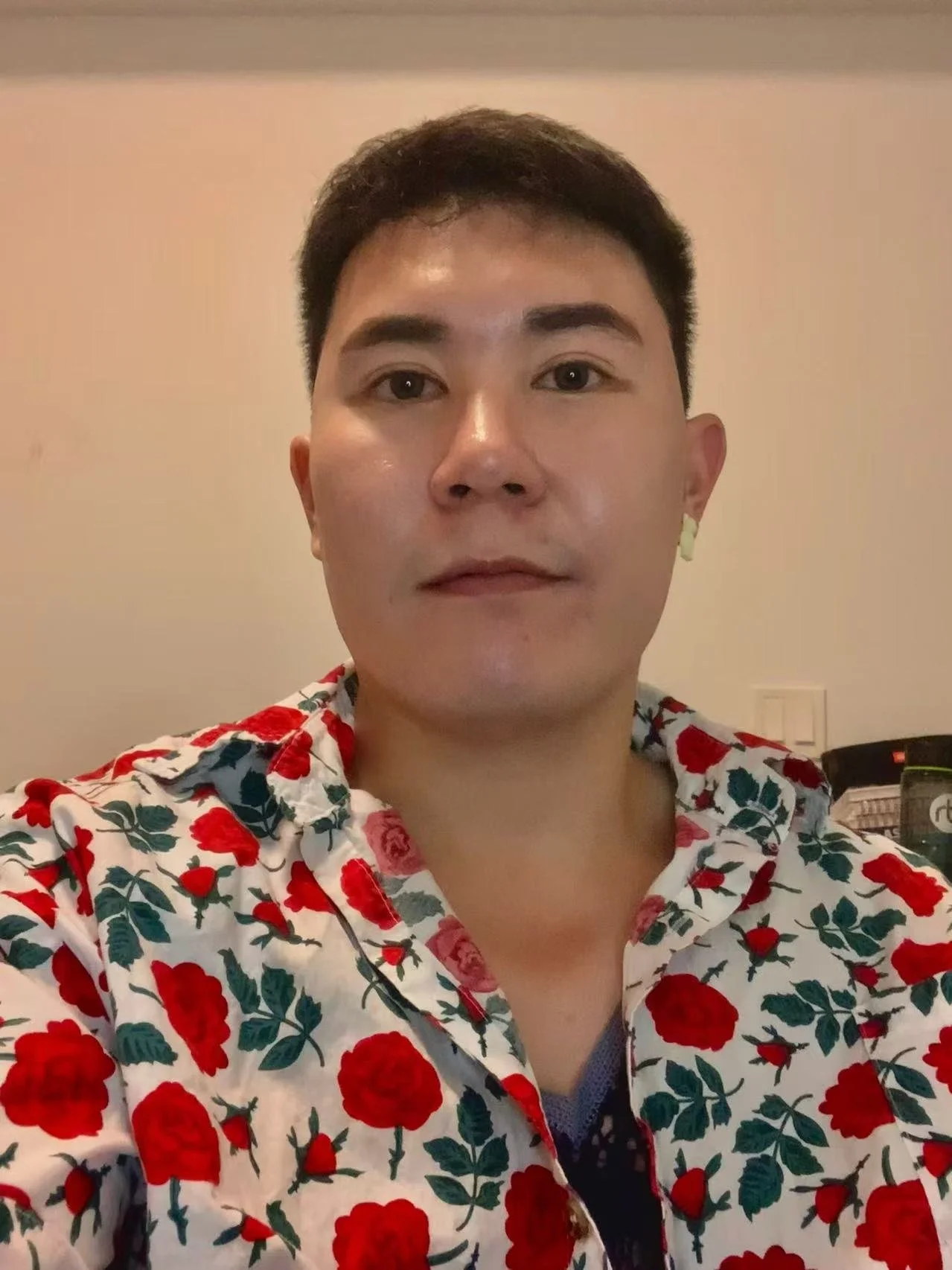 A person taking a selfie in front of a beige wall, wearing a floral shirt with red and pink flowers and dark green leaves.