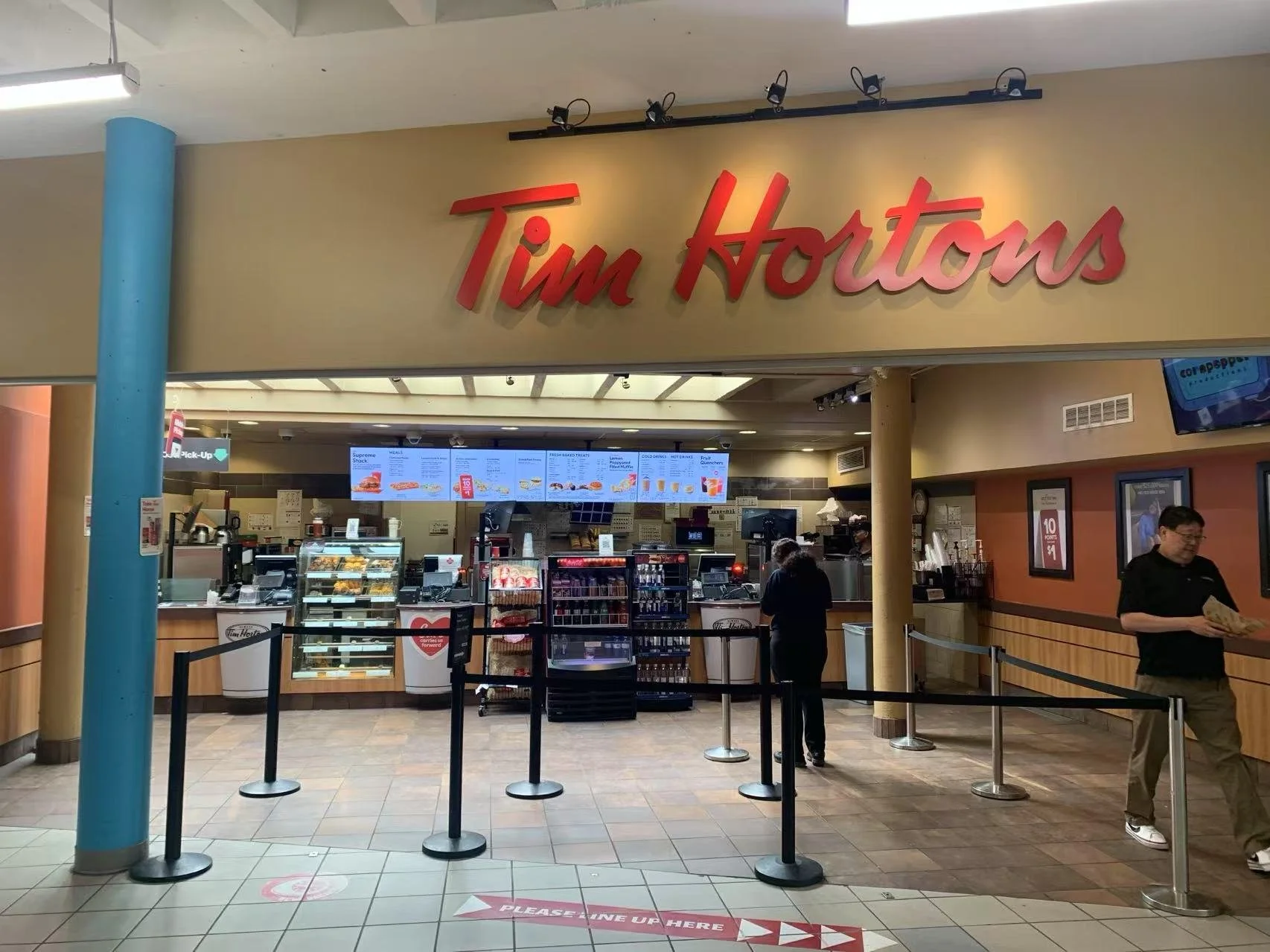 Inside a Tim Hortons coffee shop with a counter, menu digital screens, and a few customers waiting in line.