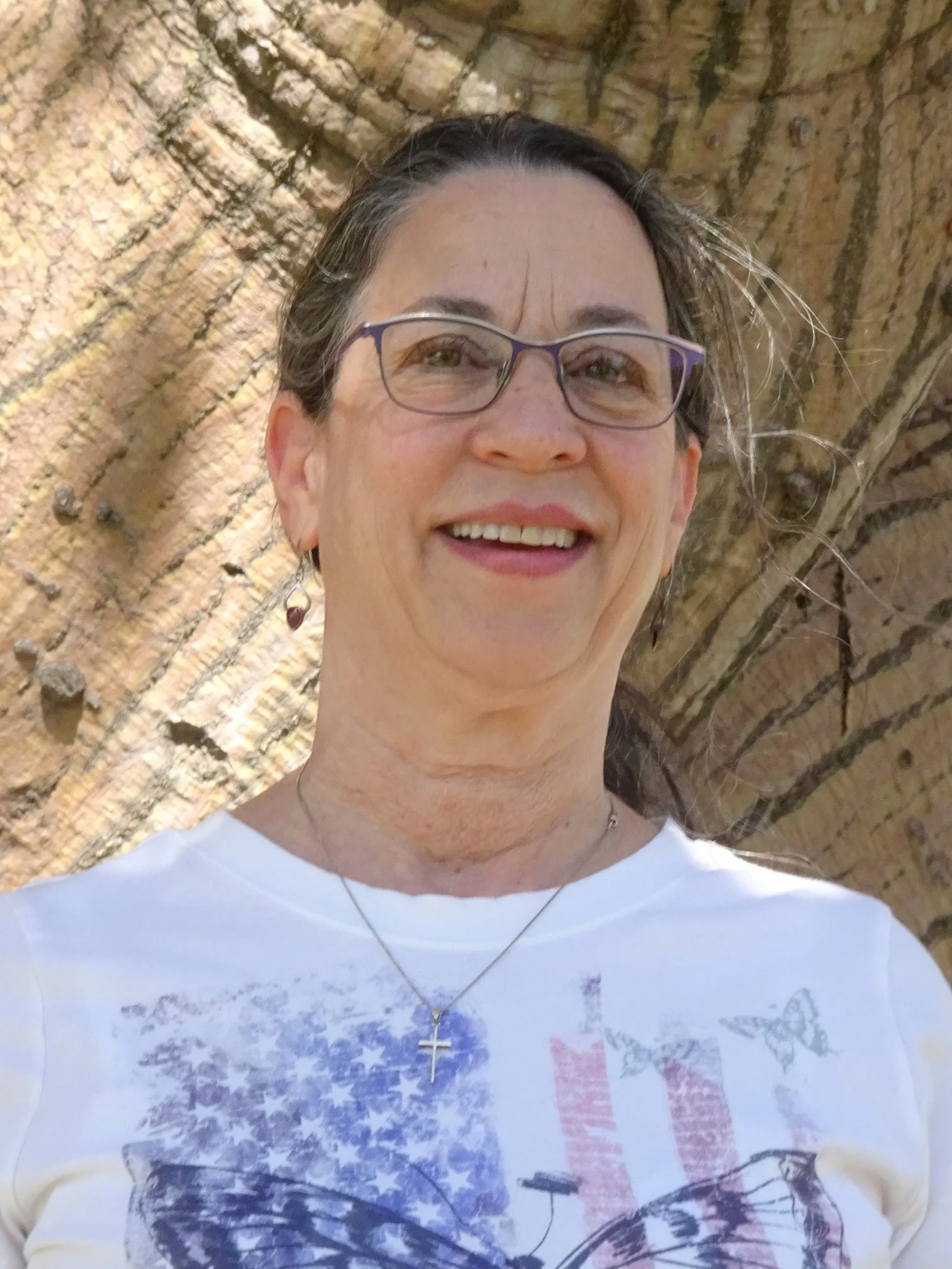 A woman with dark hair in ponytail, wearing glasses, earrings, a necklace with a cross, and a t-shirt with a graphic design, standing in front of a large tree trunk.