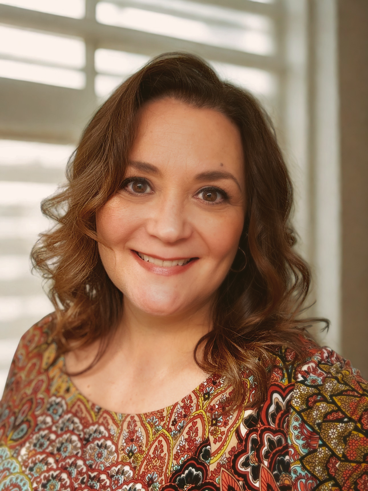 A woman with shoulder-length brown hair, smiling, wearing a colorful patterned top, with a window and blinds in the background.