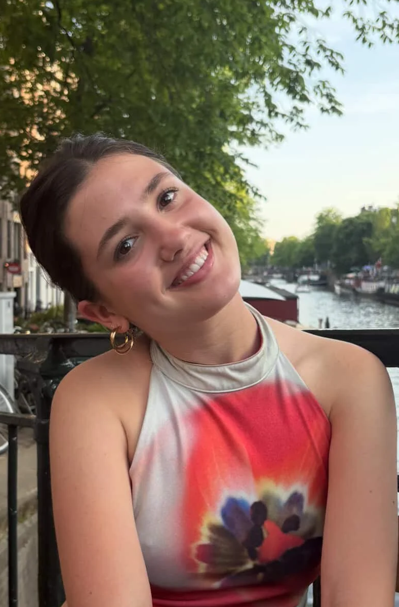 A young woman with short dark hair smiling and tilting her head, sitting outdoors by a canal with trees and boats in the background, during sunset.