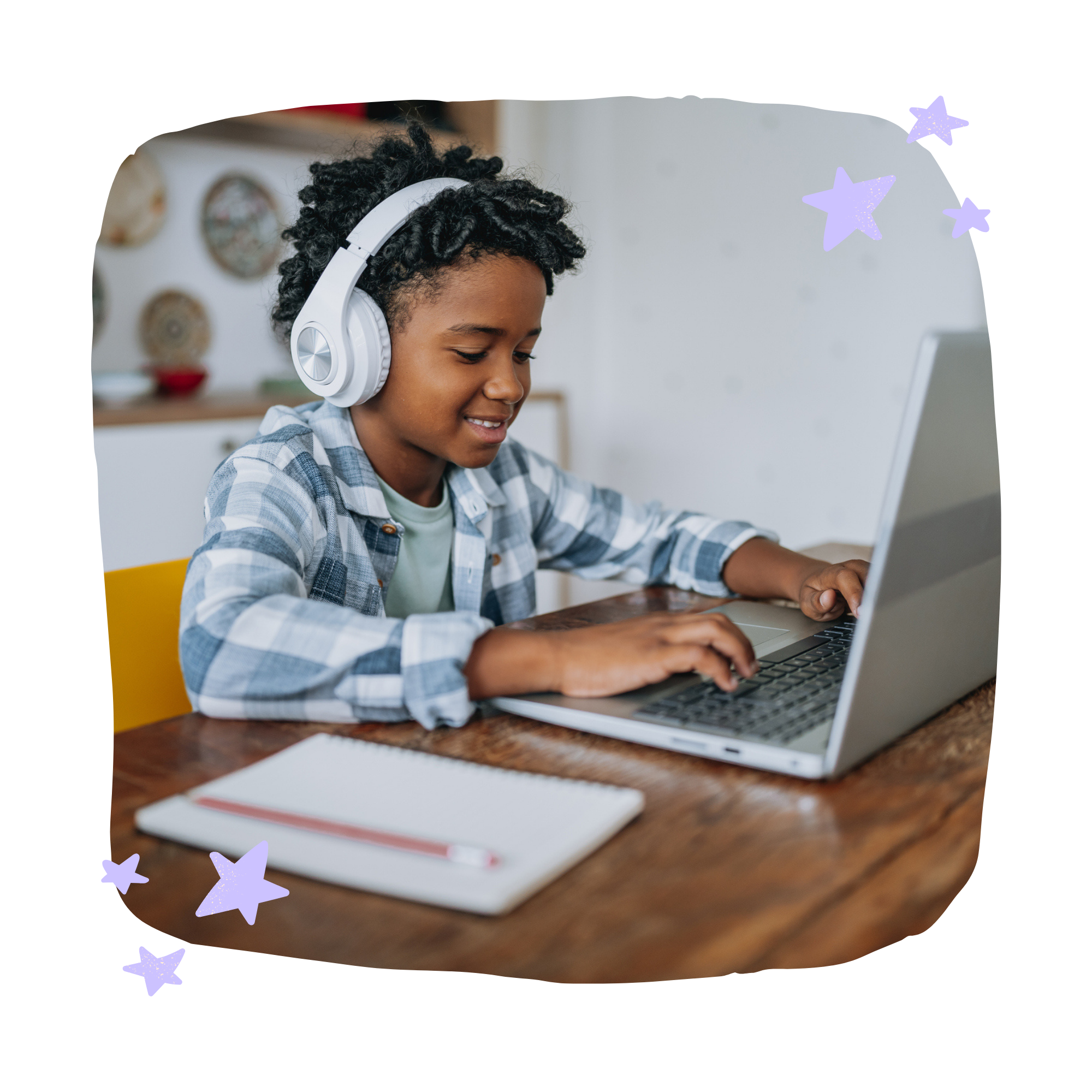 A young boy with curly hair wearing headphones and a plaid shirt, sitting at a wooden table using a laptop.