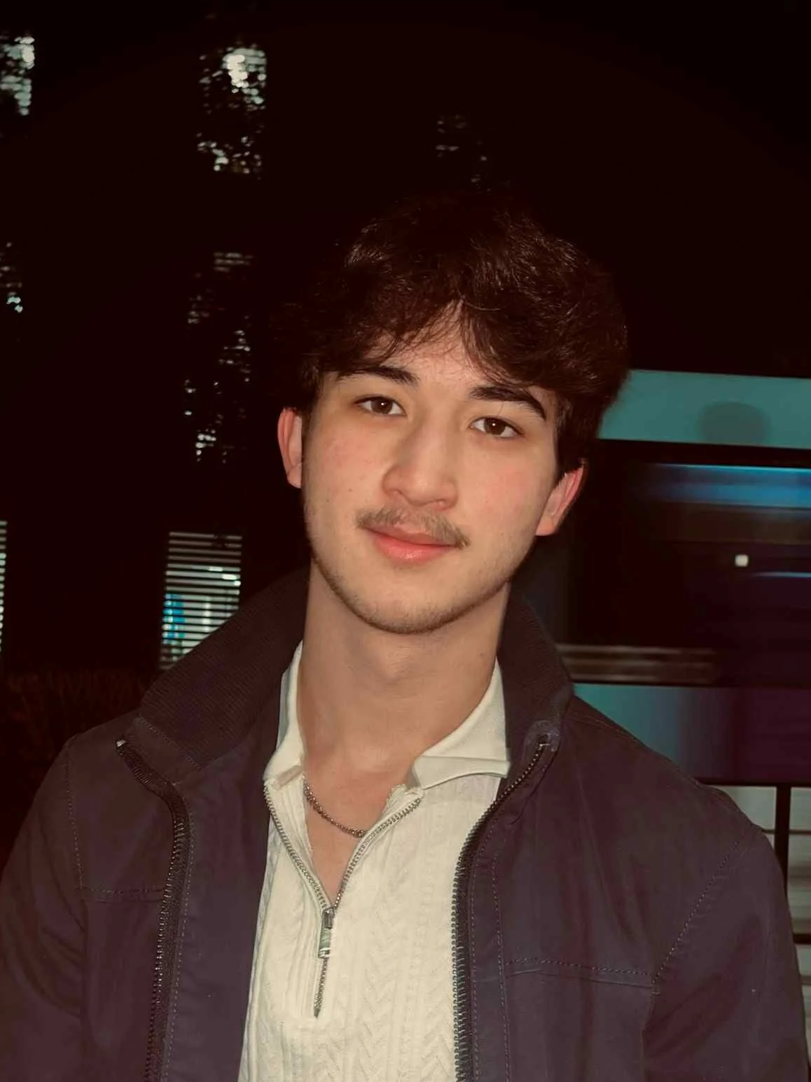 A young man with brown hair and a slight mustache smiling at the camera, wearing a beige sweater and a black jacket, standing outdoors at night with illuminated windows and a blue bus in the background.