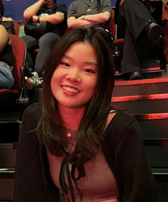 A young woman with shoulder-length dark hair and a pink top, smiling and sitting on stairs inside a theater or auditorium, with several other people seated behind her.