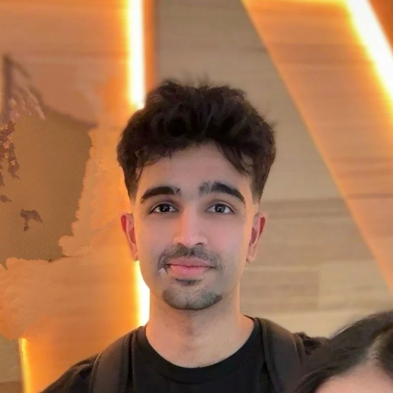 Young man with dark, styled hair and facial hair smiling at the camera in an indoor setting with warm lighting and a blurred background.