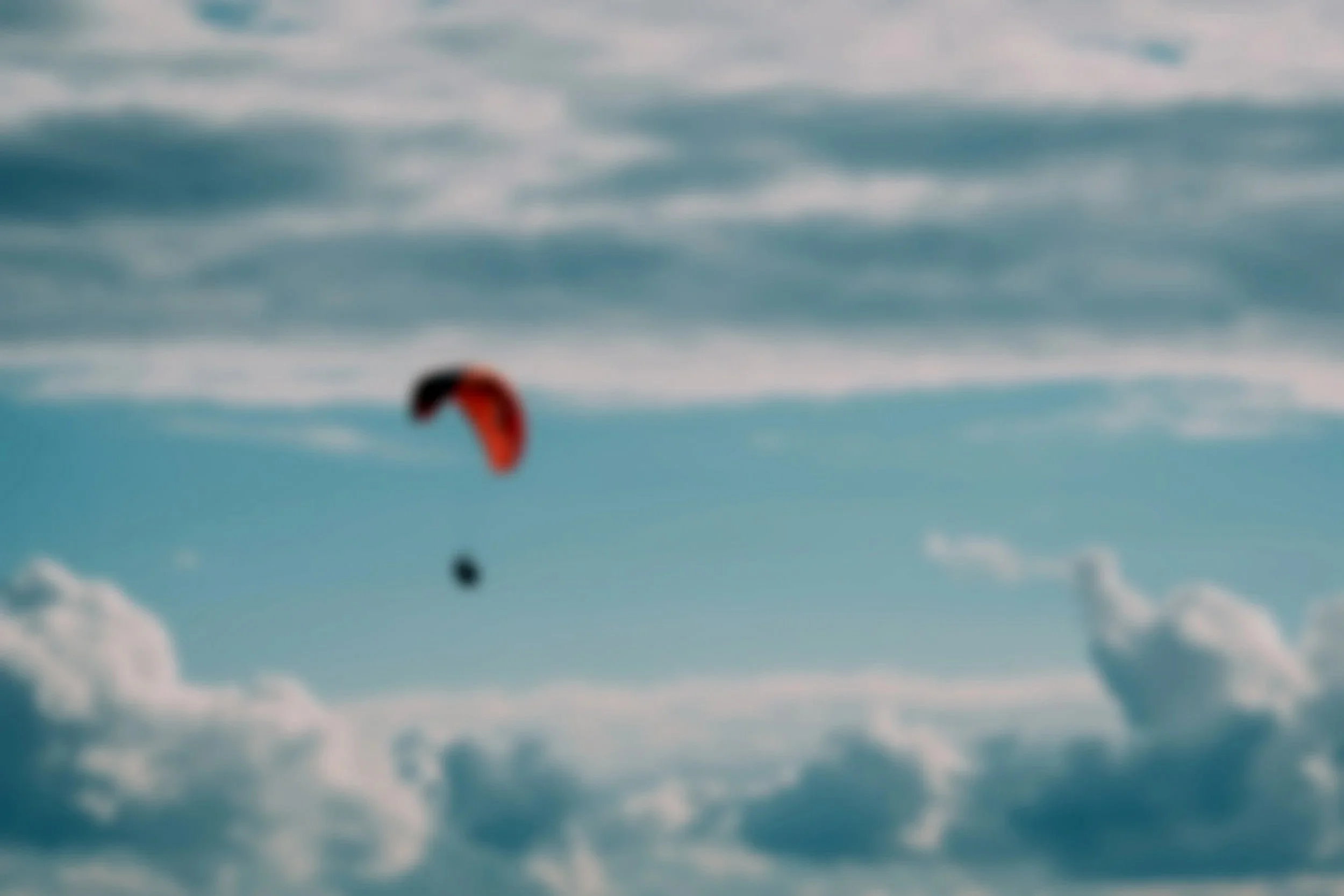 A blurred image of a person paragliding over snowy terrain under a partly cloudy sky.