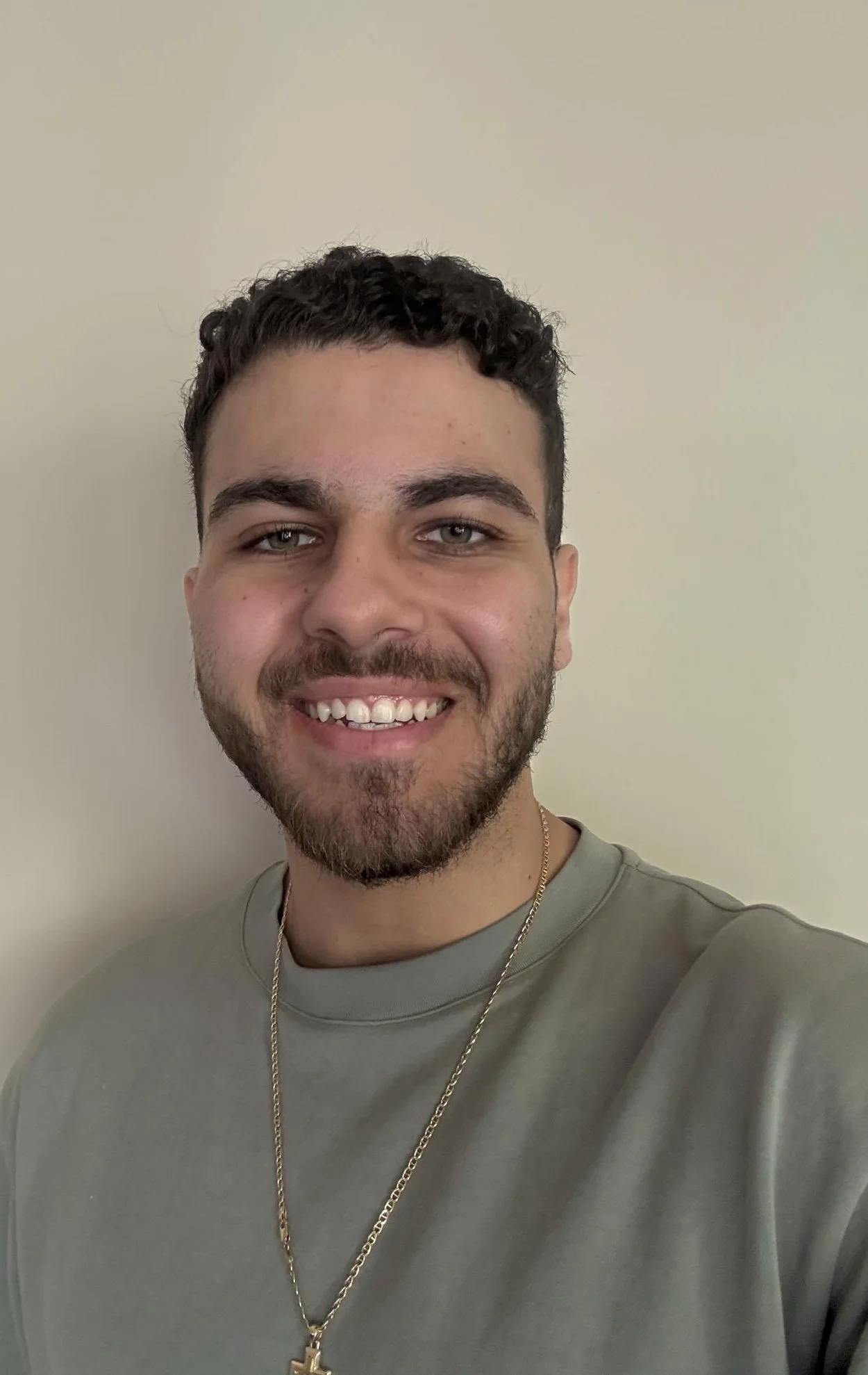 A young man with short curly dark hair, a beard, and light skin smiling and wearing a gray shirt and a gold chain with a cross pendant, standing against a plain beige wall.