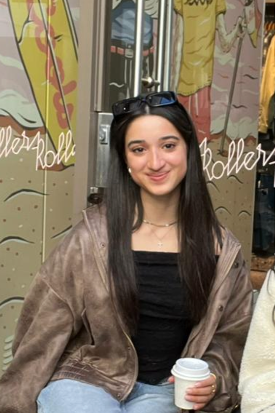 A young woman with long dark hair sitting at a table in a cafe or restaurant, holding a white disposable coffee cup with a lid, smiling at the camera. She is wearing a brown jacket, black top, and has sunglasses on her head.