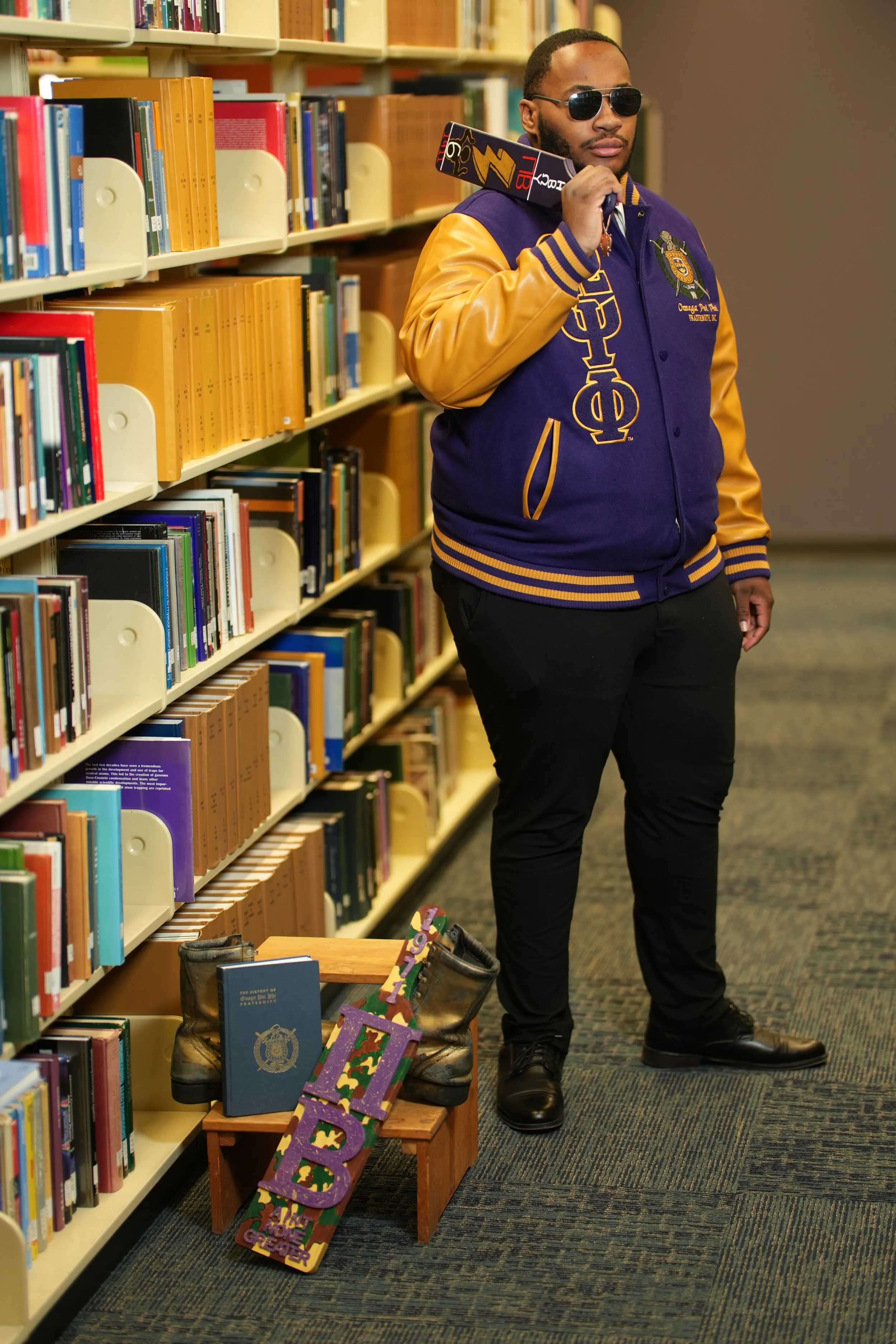 A man in a purple and yellow fraternity jacket and sunglasses standing in a library aisle, holding a book to his ear with a serious expression.
