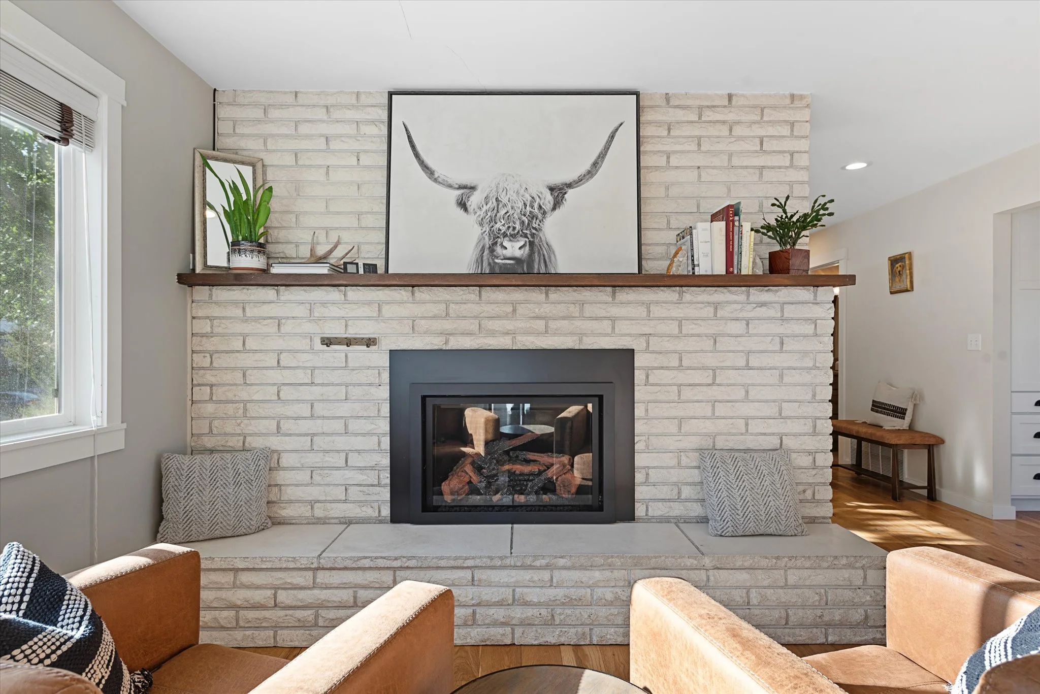 Living room with a white brick fireplace, black frame, and a large black and white highland cow painting above it. Decor includes potted plants, books, and cushions, with a window to the left.