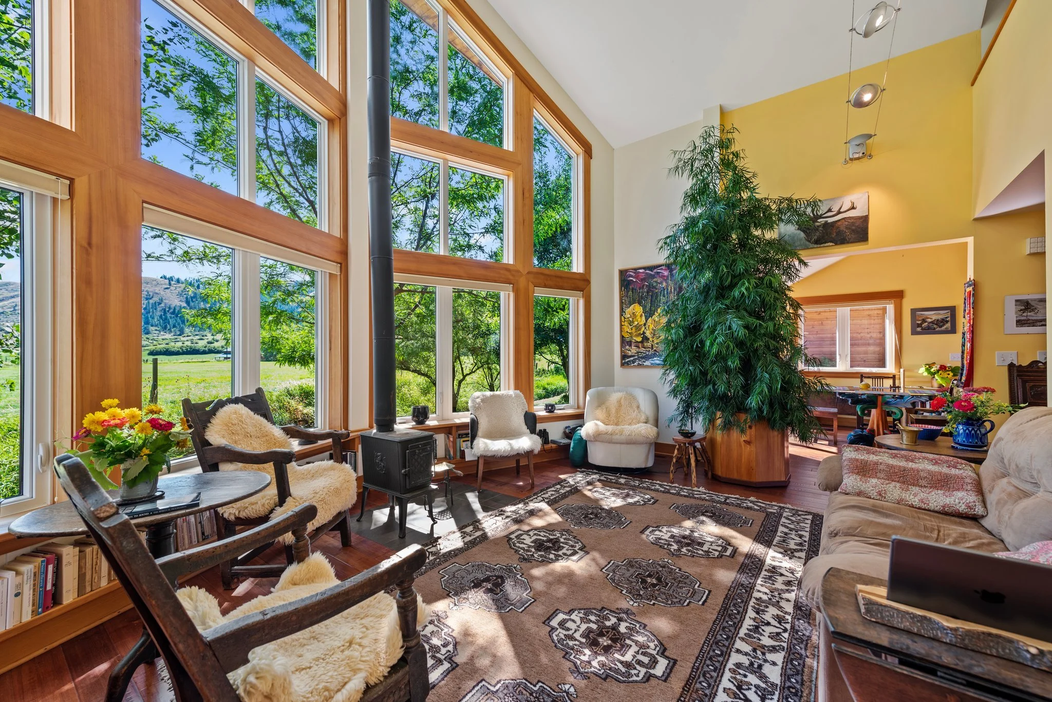 Living room with large windows, wooden floors, and cozy furniture, including a sofa, chairs with fur throws, a fireplace, and decorative plants.