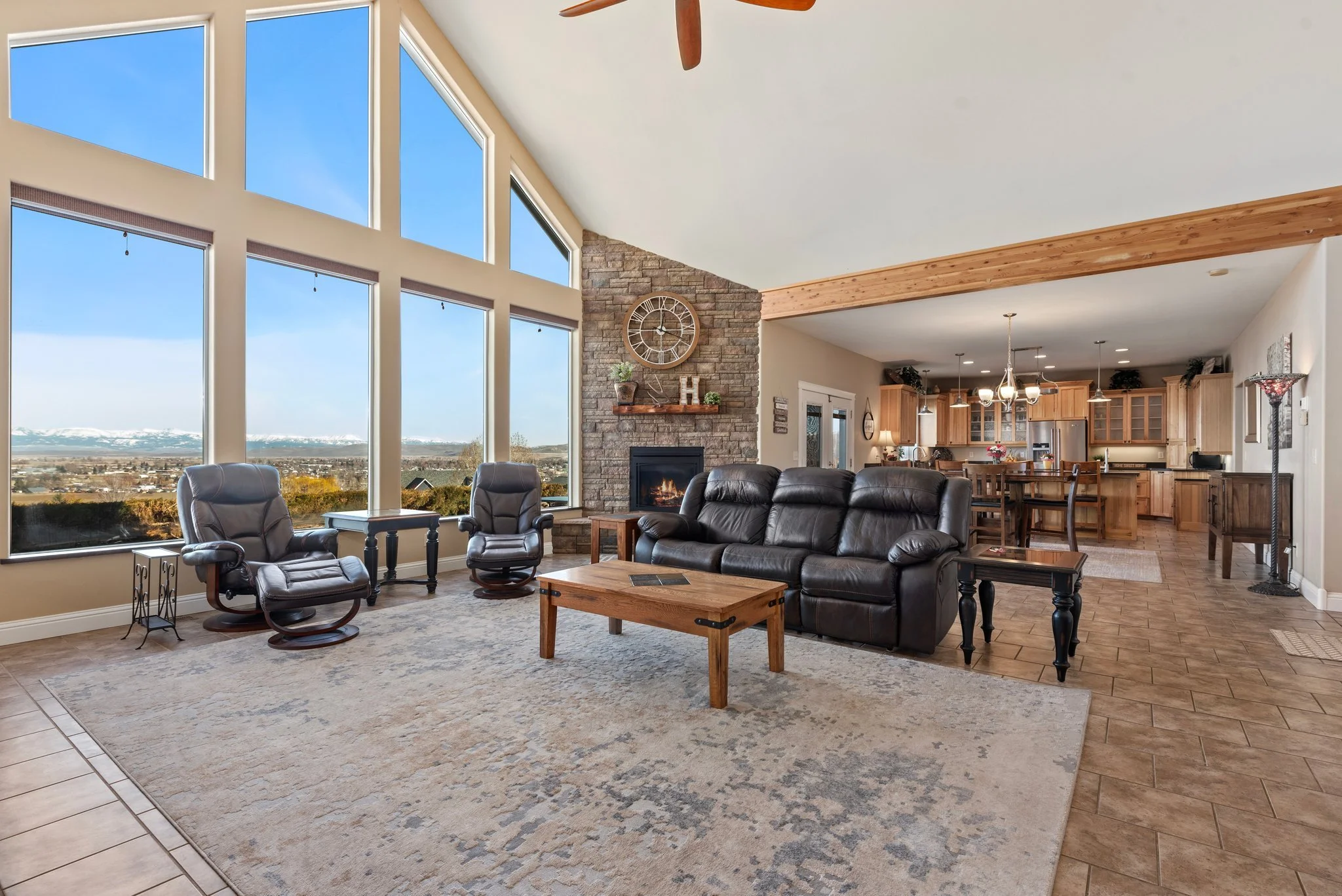 Living room with large windows showing snowy mountains, a stone fireplace, leather sofas, armchairs, and a wooden coffee table.