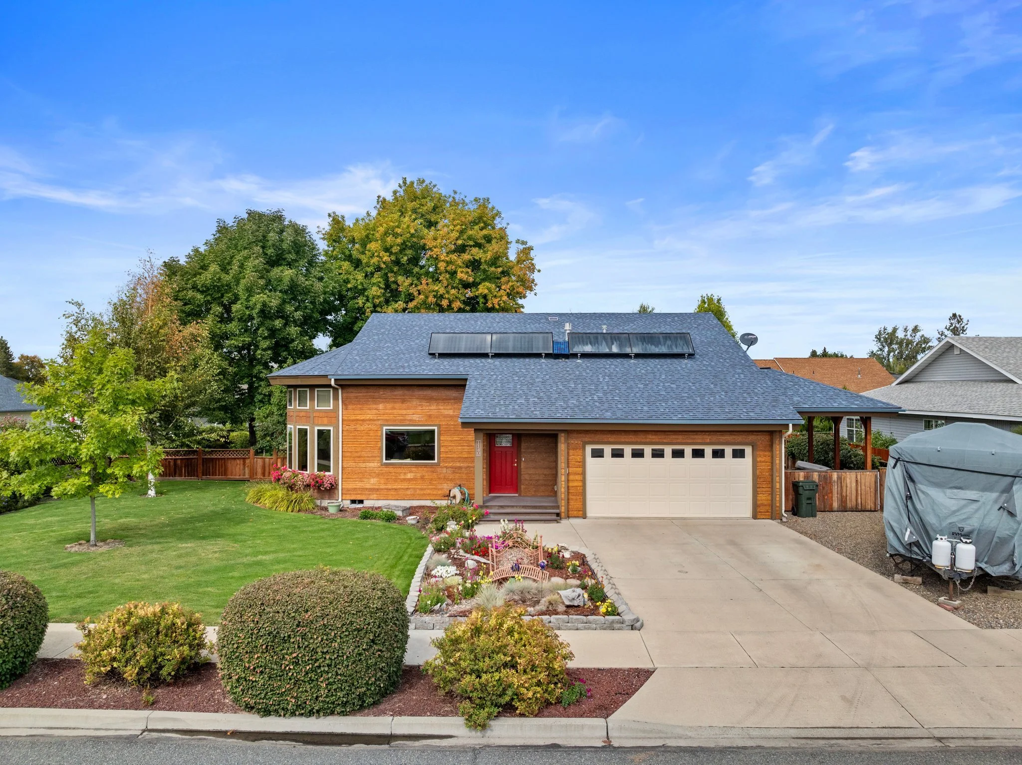 A modern two-story house with solar panels on the roof, a red front door, a manicured front yard with shrubs and flowers, a concrete driveway, and neighboring houses in a suburban neighborhood.