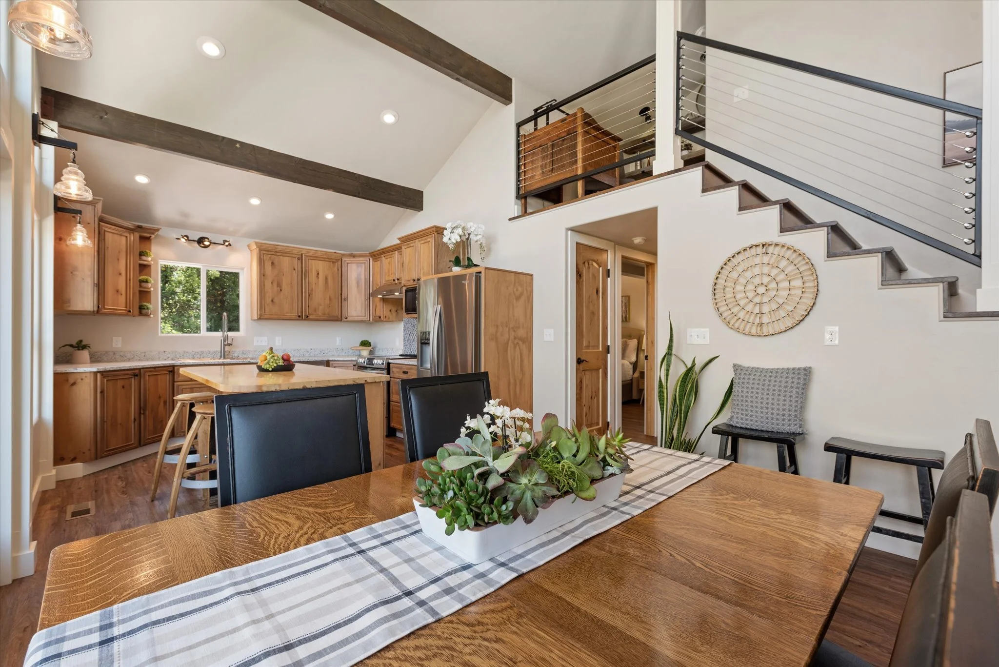 Open-concept kitchen and dining area with wooden cabinets, stainless steel refrigerator, wooden dining table with black chairs, decorative plant centerpiece, staircase with modern railing, and cozy seating nook in the background.