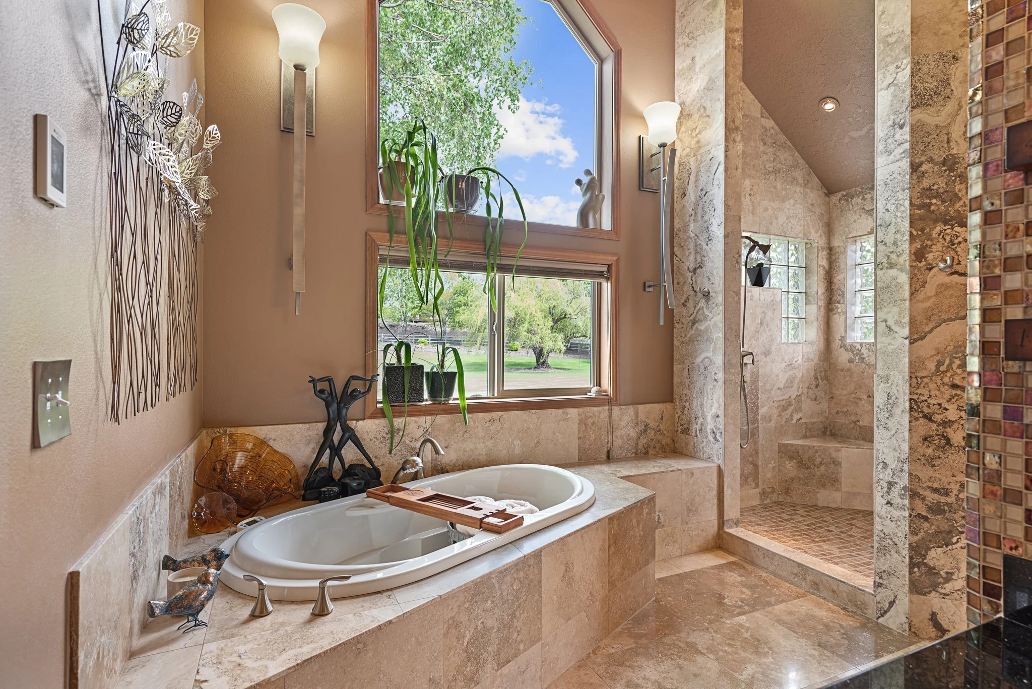 Luxury bathroom with a bathtub and a walk-in shower, beige marble tiles, two tall lights, and large windows overlooking a green outdoor scene.