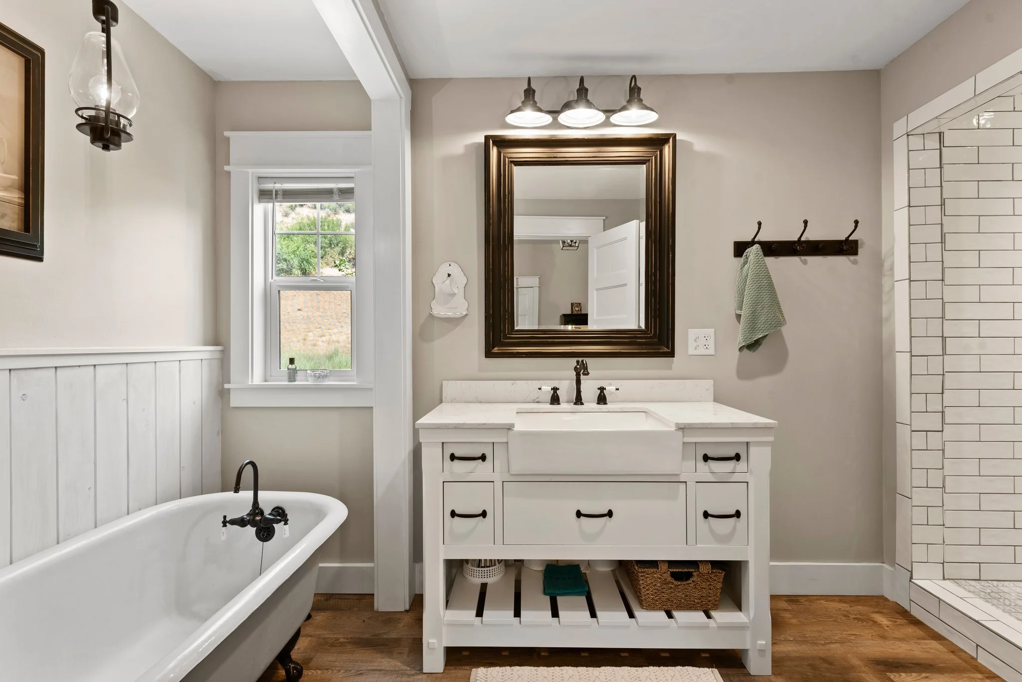 Bathroom with a white freestanding bathtub, a window, a white vanity with a sink, a large framed mirror, a wall-mounted light fixture, a towel rack with a green towel, and a tiled shower stall.