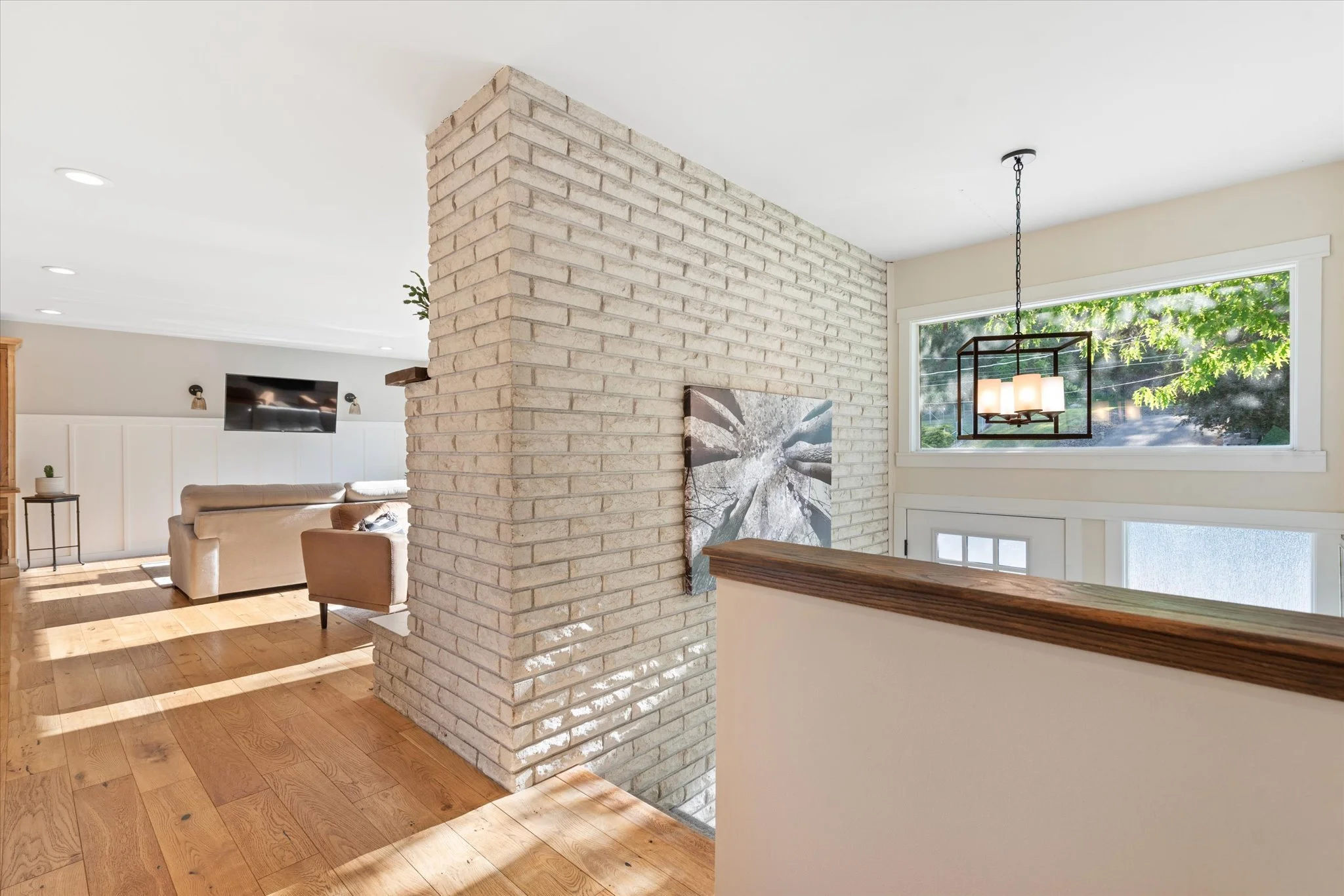 Living room with a brick partition wall, wood flooring, beige couches, wall-mounted television, and a window with green foliage outside.
