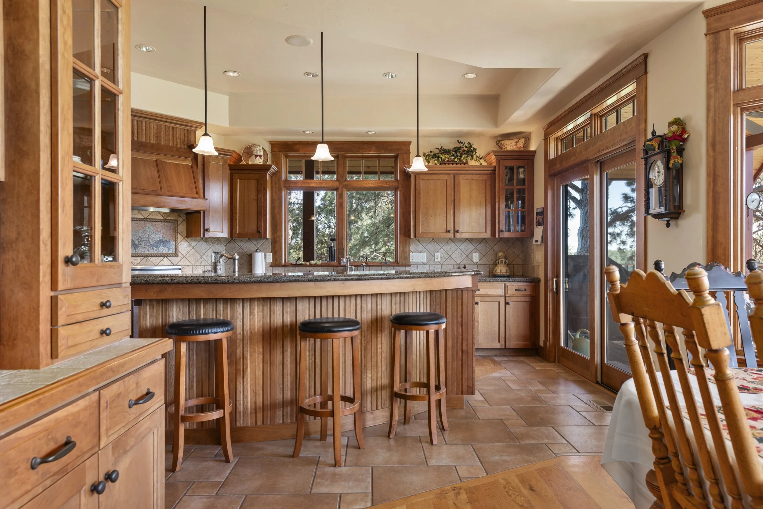 A cozy kitchen with wooden cabinets, a granite countertop, and a breakfast bar with three black-stool seats. There are pendant lights hanging above the breakfast bar, and a window above the sink overlooking trees outside. To the right, there is a sli
