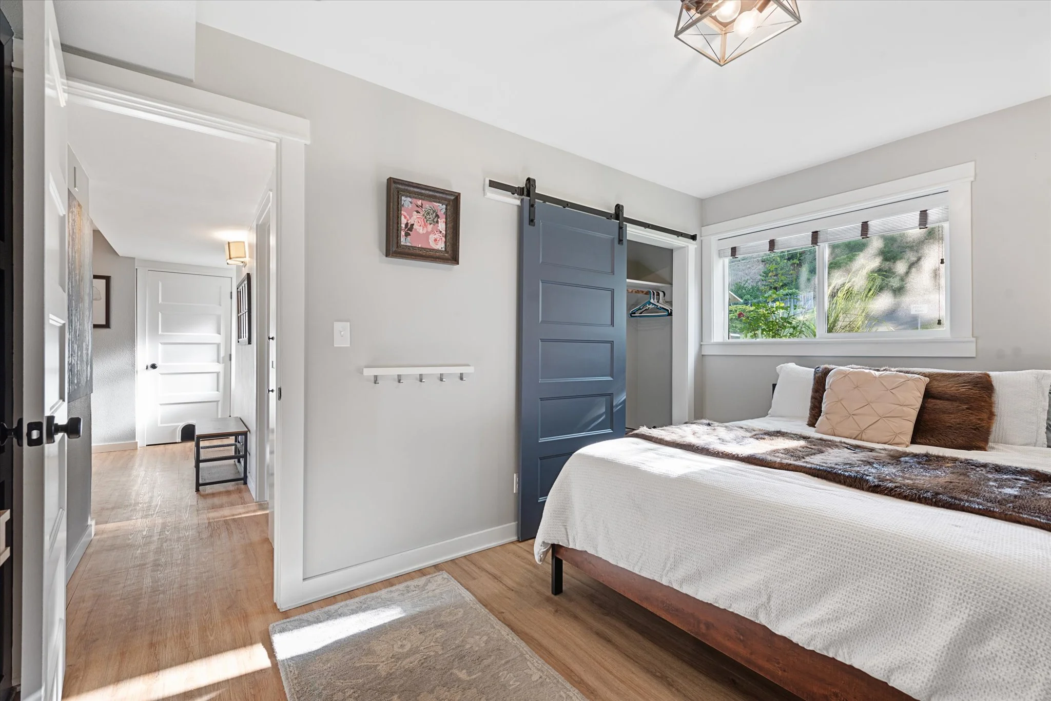 A bedroom with a bed covered in white bedding, accent pillows, and a fur throw, a large window with blinds, a sliding closet door, and a hallway with hardwood flooring.