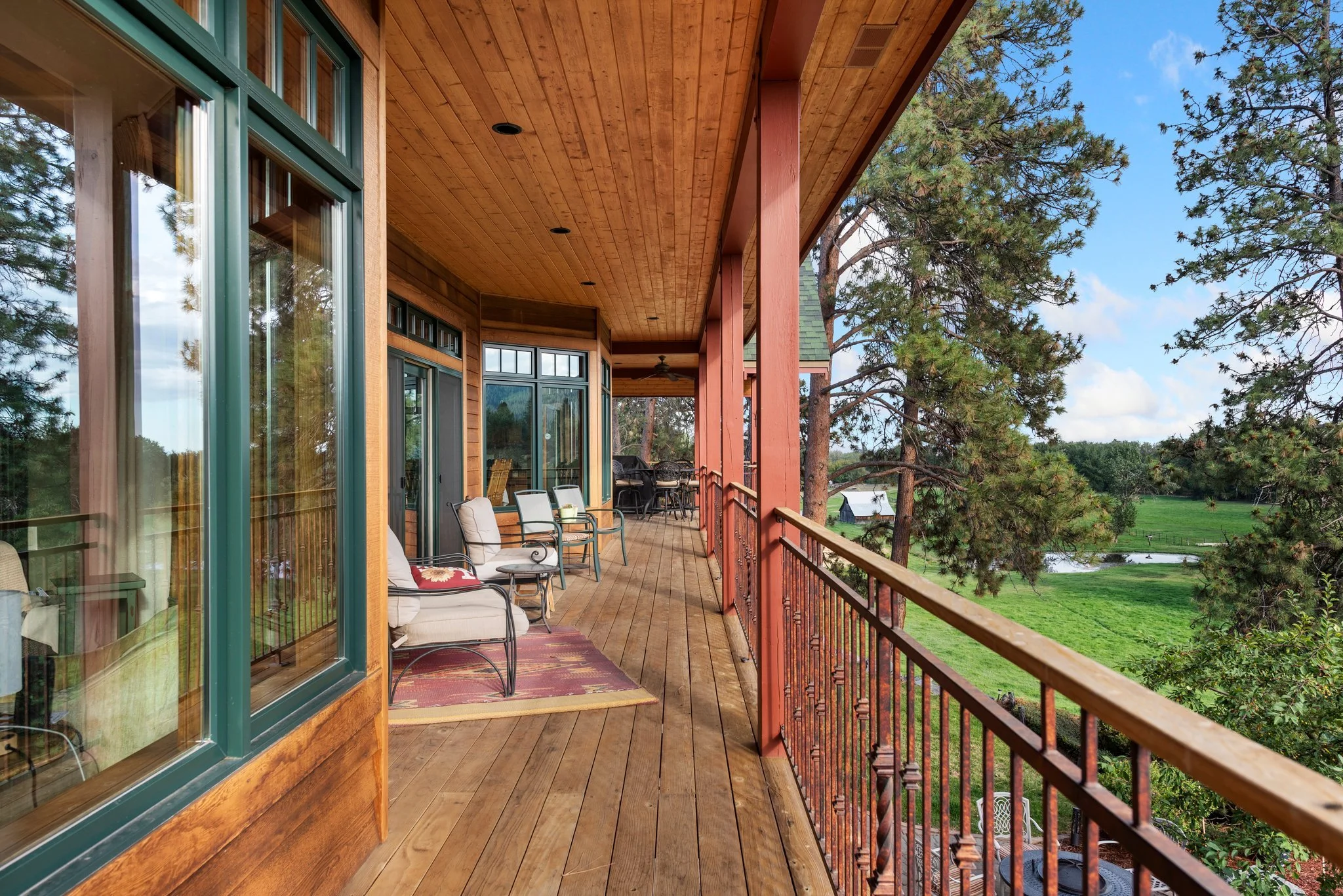 A spacious wooden balcony with outdoor furniture, including chairs with cushions, overlooking a green landscape with trees, a small pond, and a barn in the distance.