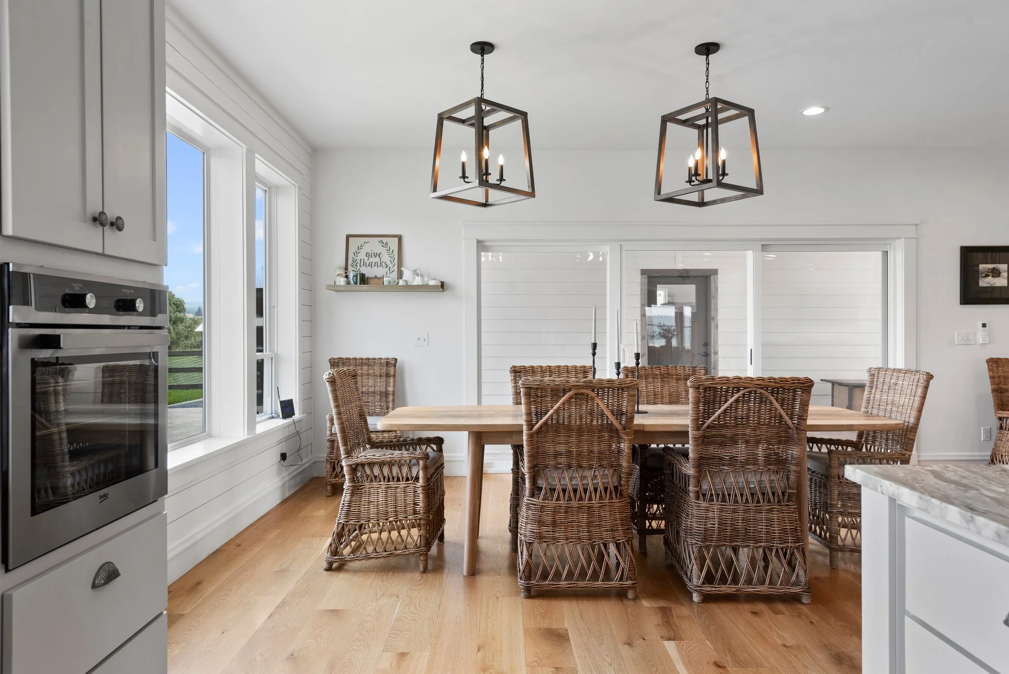 Dining room with a wooden table, eight wicker chairs, modern black metal light fixtures, white walls, and a large sliding glass door.