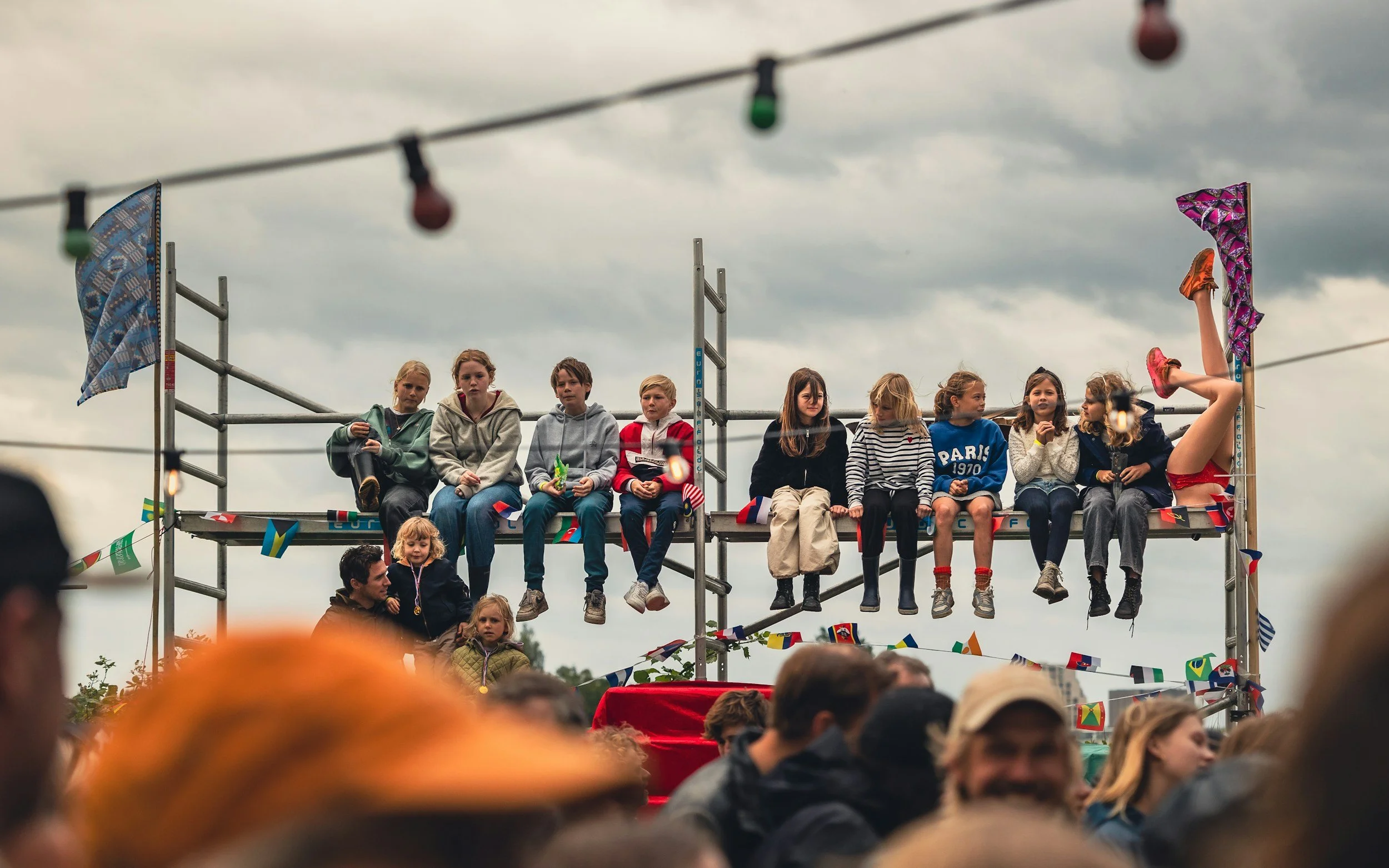 Children sitting on a high platform at an outdoor event, with flags and colorful banners hanging, and a crowd in the foreground, under a cloudy sky.