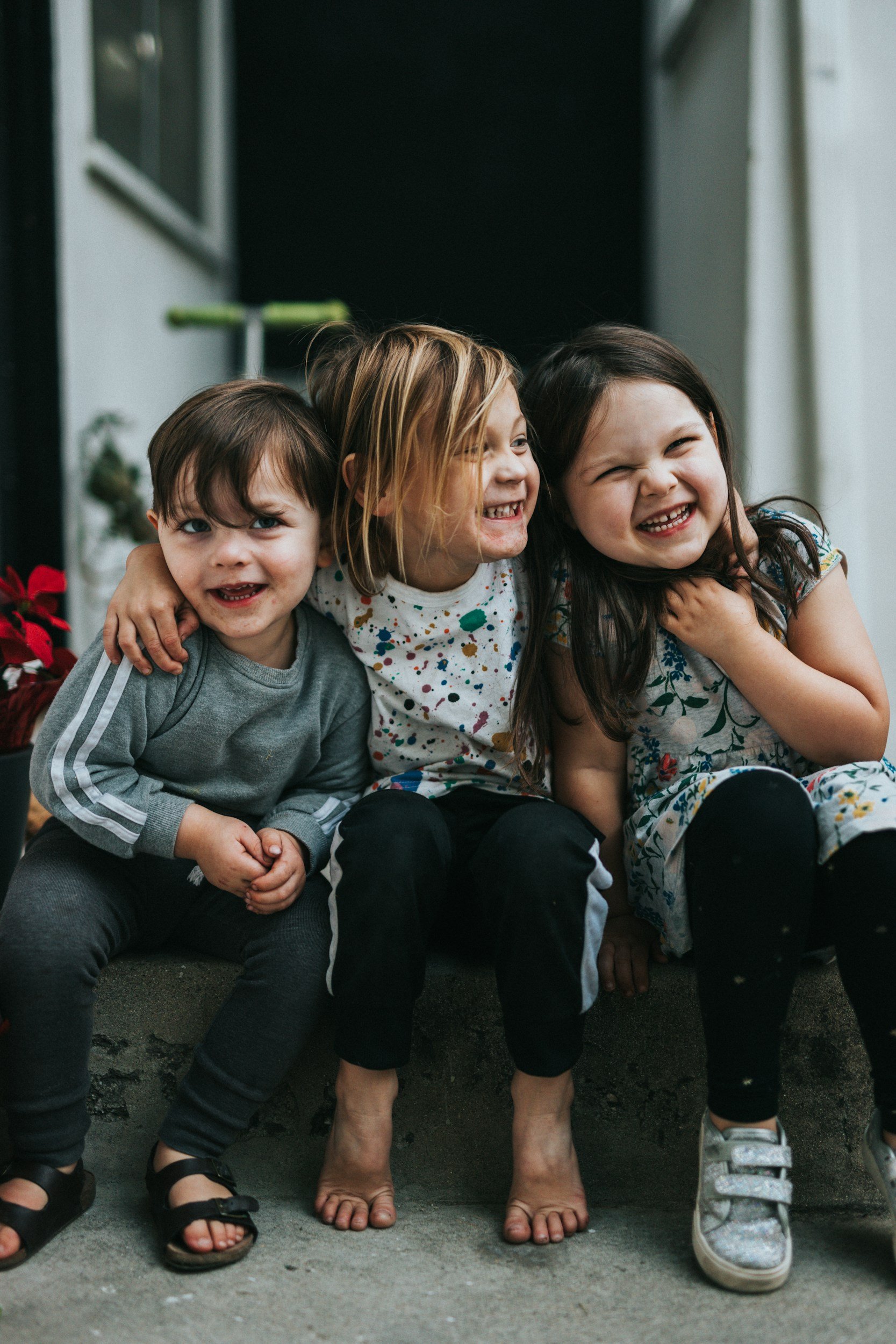 Three young children sitting close together on a concrete surface in front of a house, smiling and laughing, with one child being supported by the others.