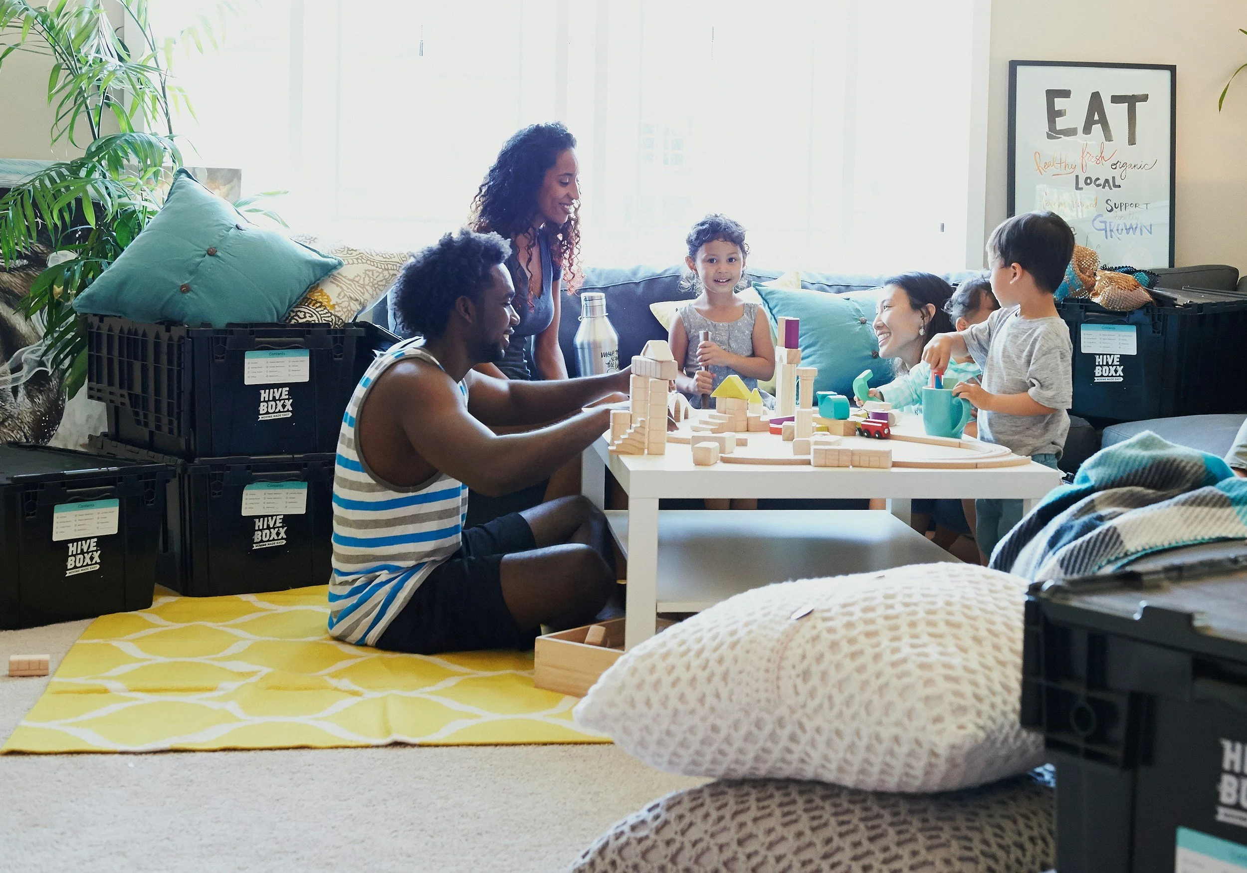 A diverse group of children and adults playing with wooden building blocks at a table in a bright living room. There are storage crates labeled "Hive Boxx" and a framed poster with the word "Eat" on the wall.