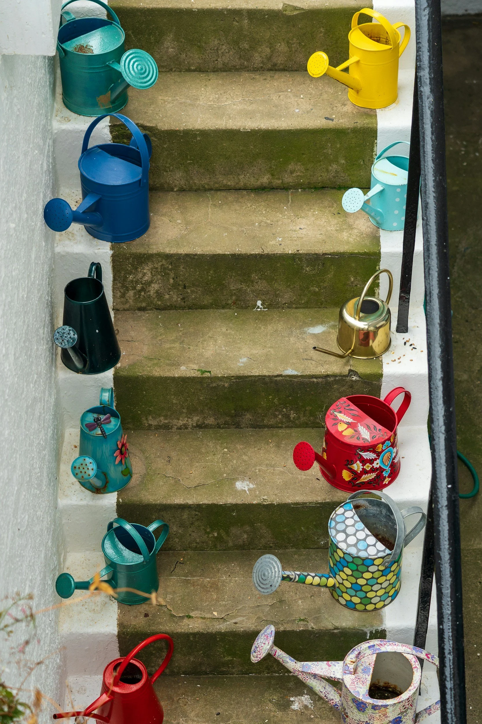 Colorful watering cans placed on outdoor concrete stairs, with moss and dirt on the steps, and a black metal handrail on the right side.