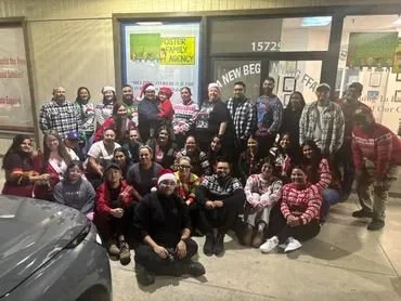 Group of people, some wearing Christmas-themed attire, gathered in a parking lot in front of a building with signs, smiling and posing for a photo.