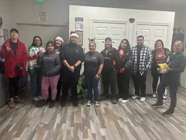 Group of twelve people standing indoors, some wearing holiday attire like Santa hats and Christmas sweaters, in front of closed white doors.