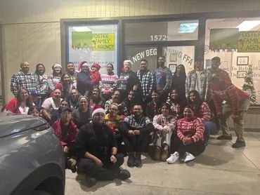 Group of people gathering indoors, some wearing Santa hats, with banners and signs in the background.