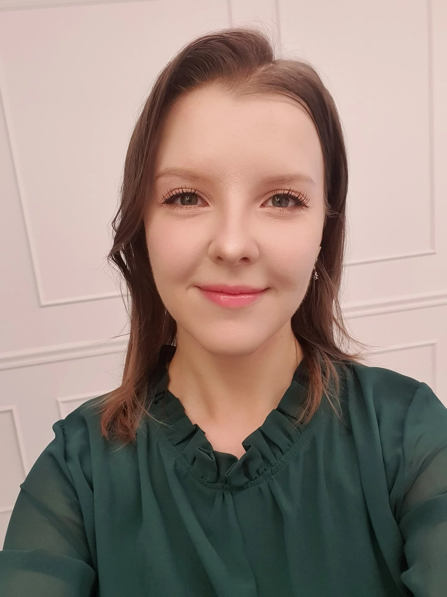 A young woman with shoulder-length brown hair, smiling, wearing a dark green blouse, standing in front of a light-colored paneled wall. Coach Dina. soaring with Dina.