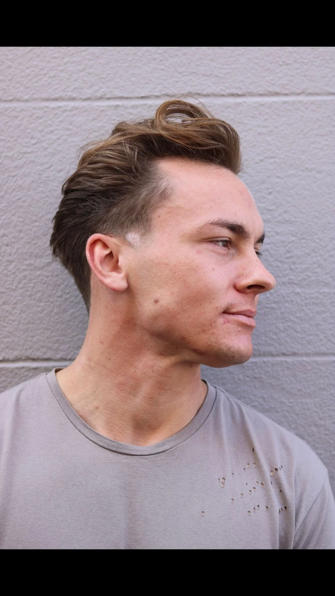 Side profile of a young man with styled brown hair, wearing a gray T-shirt with small holes, standing against a textured gray wall.