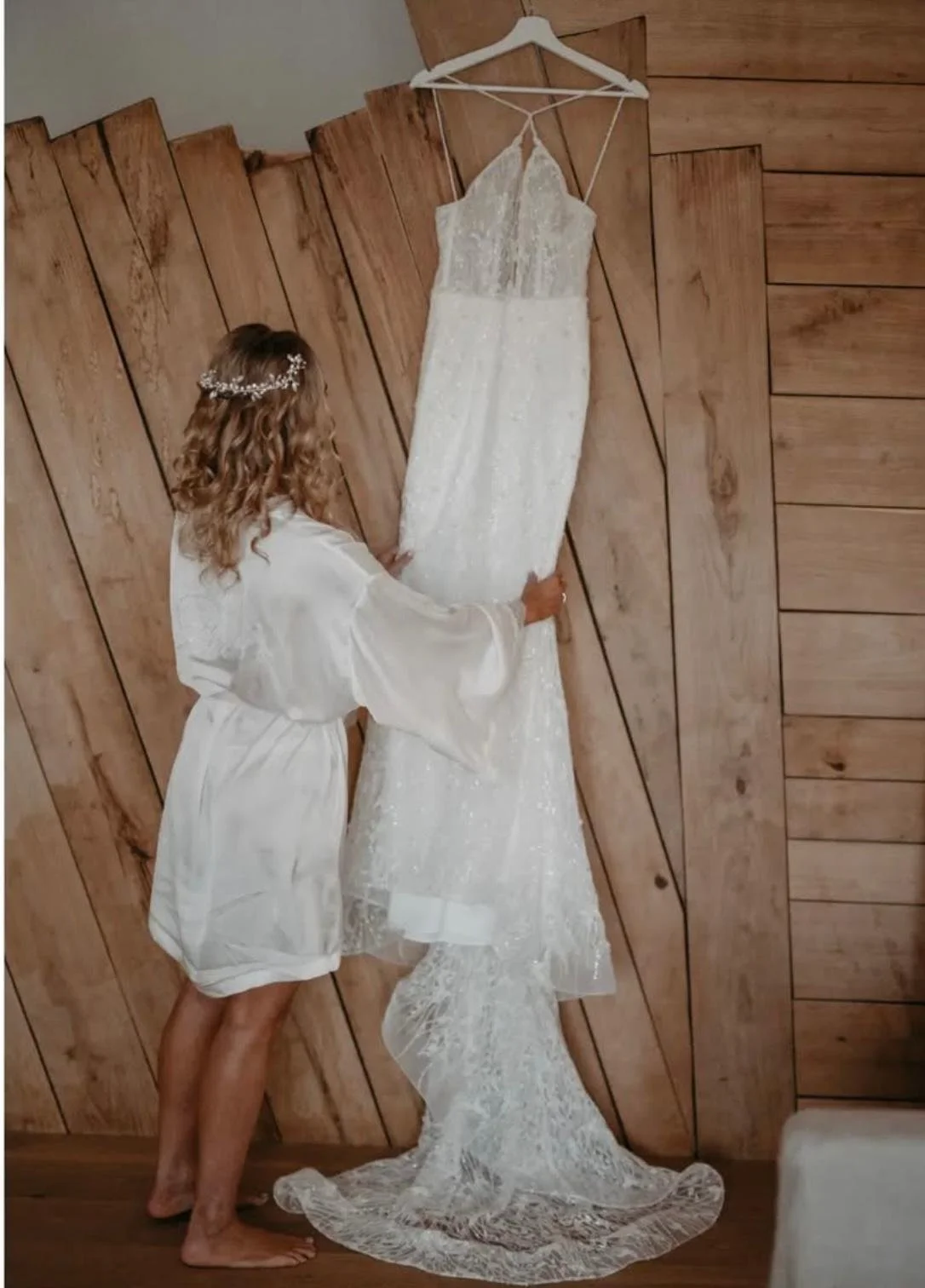Une femme portant un peignoir blanc regarde une robe de mariée suspendue au mur en bois.